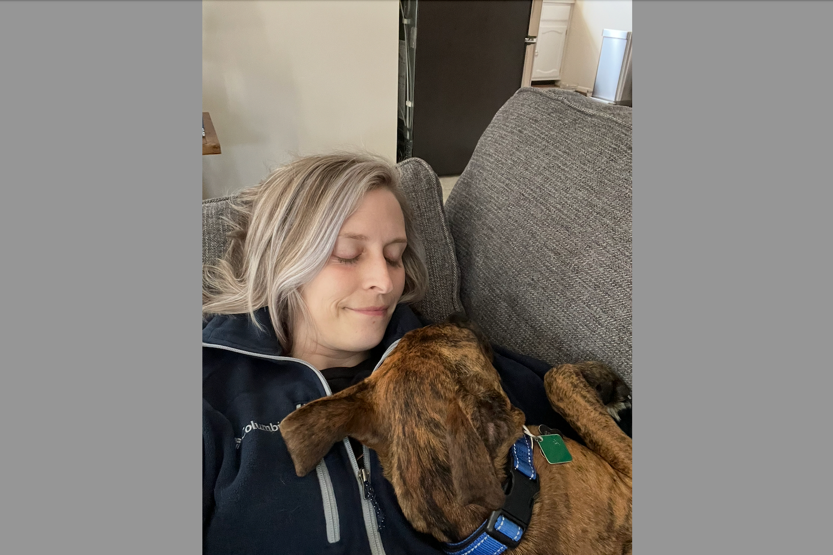 Woman with blond hair resting on a gray sofa, eyes closed, cuddling a brindle dog wearing a blue collar in a living room.",
