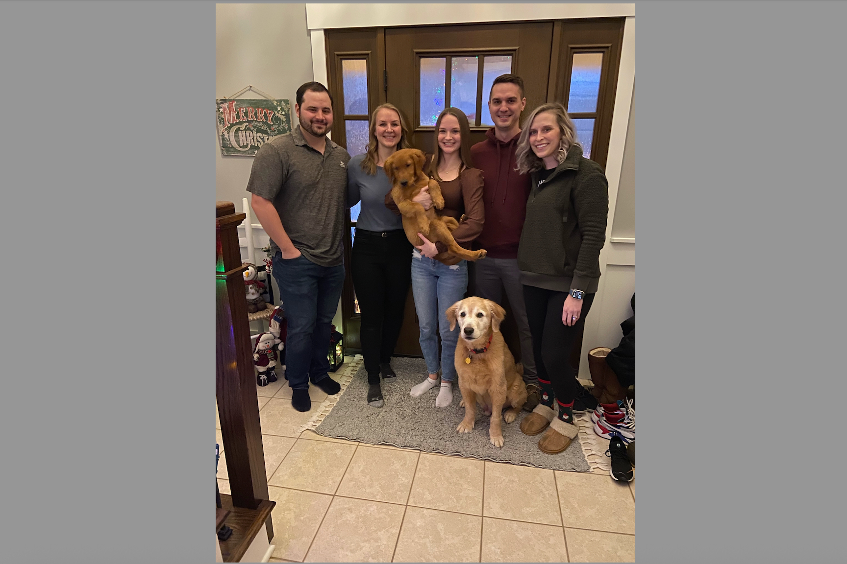 Six adults posing together in a home entryway with a puppy held aloft and a golden retriever seated on the floor.