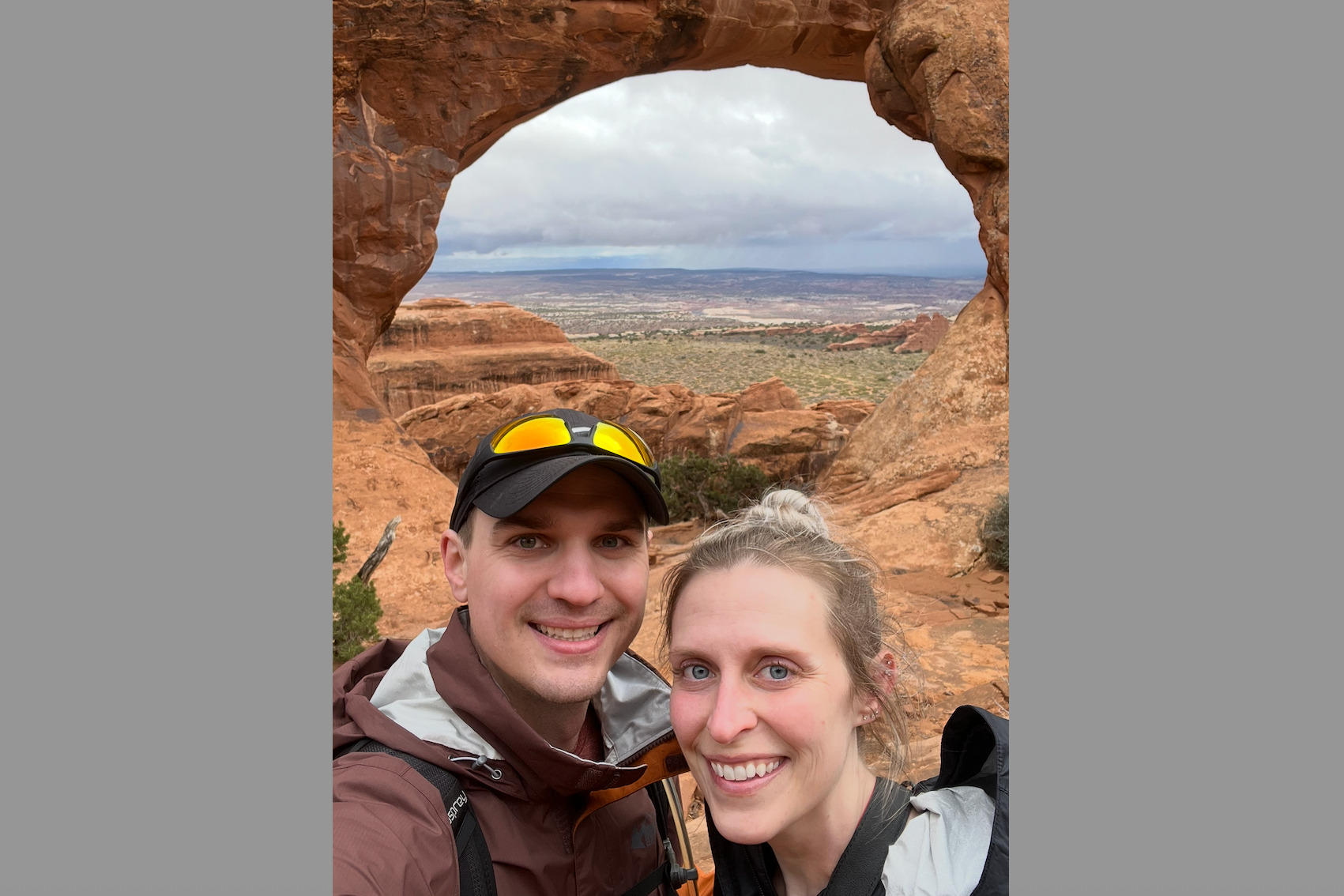 Smiling couple taking a selfie beneath a large red rock arch, with a desert valley in the distance.