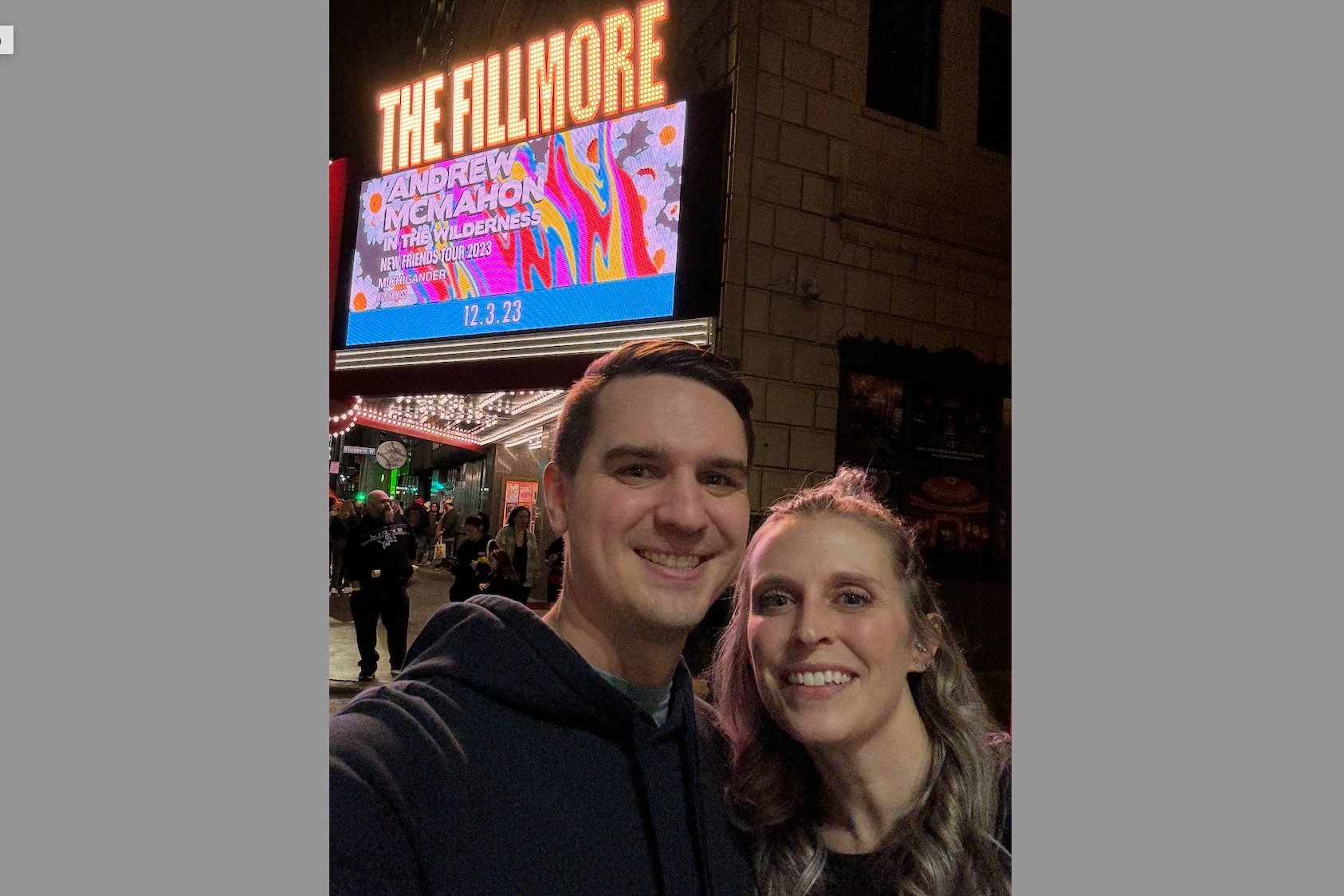 Selfie of a smiling couple in front of The Fillmore marquee at night, with a colorful concert sign in the background