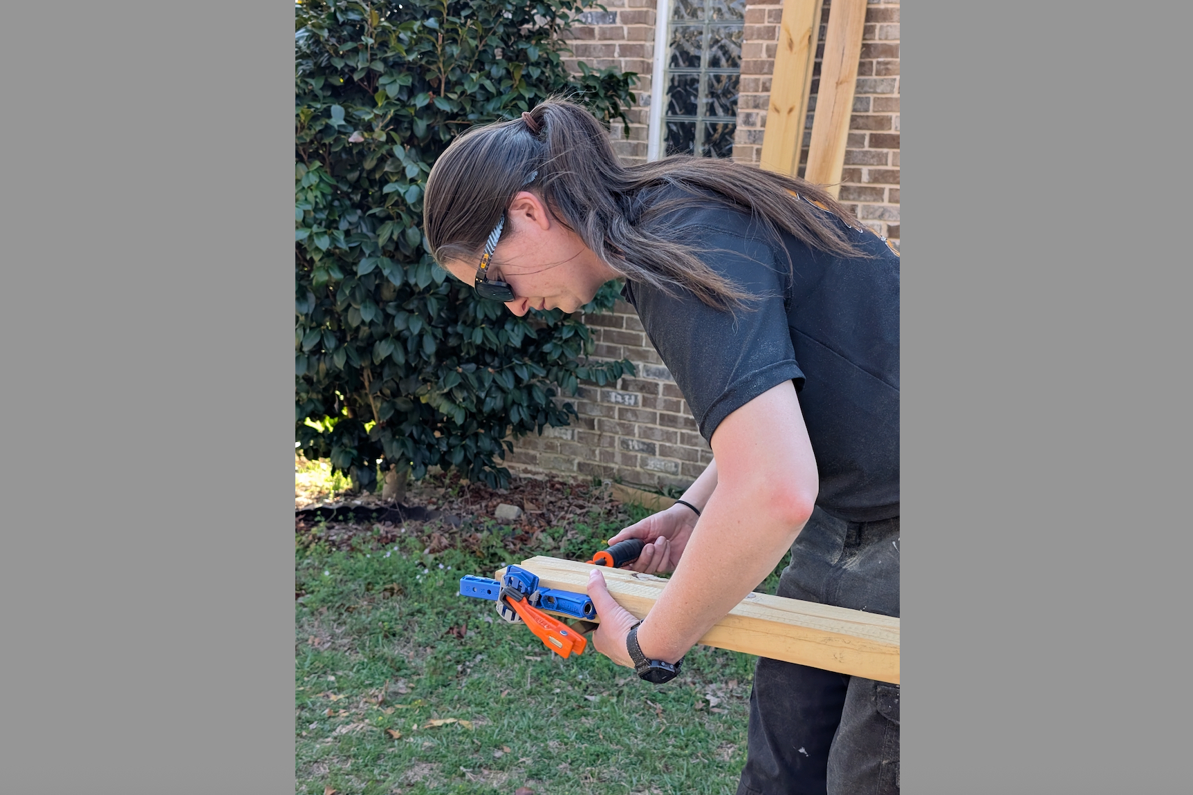 Person wearing safety glasses clamps a wooden board outdoors with a power tool in use near a brick house.