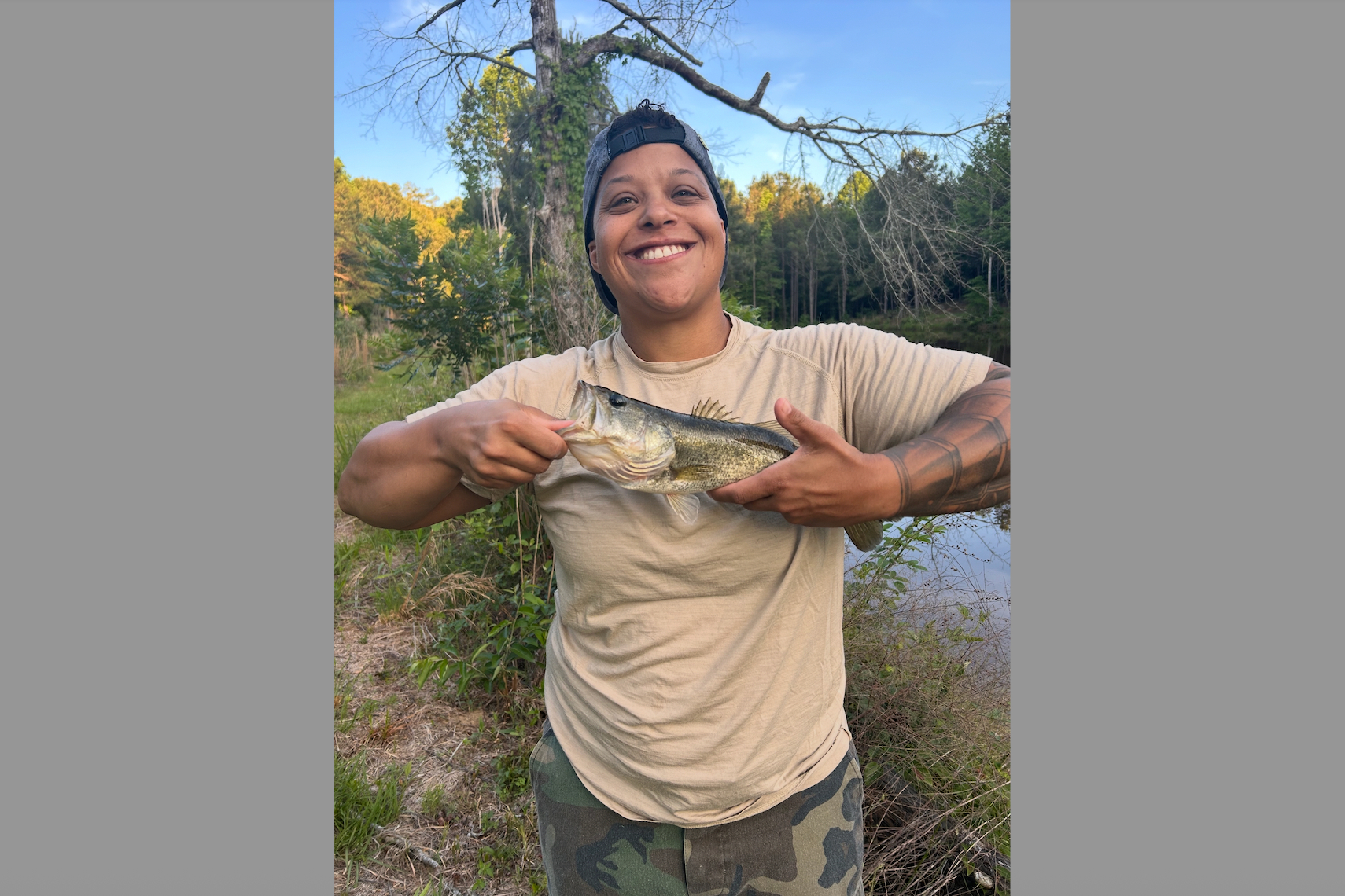 Person smiling while holding a largemouth bass outdoors by a pond, wearing a beige T-shirt and camo pants.