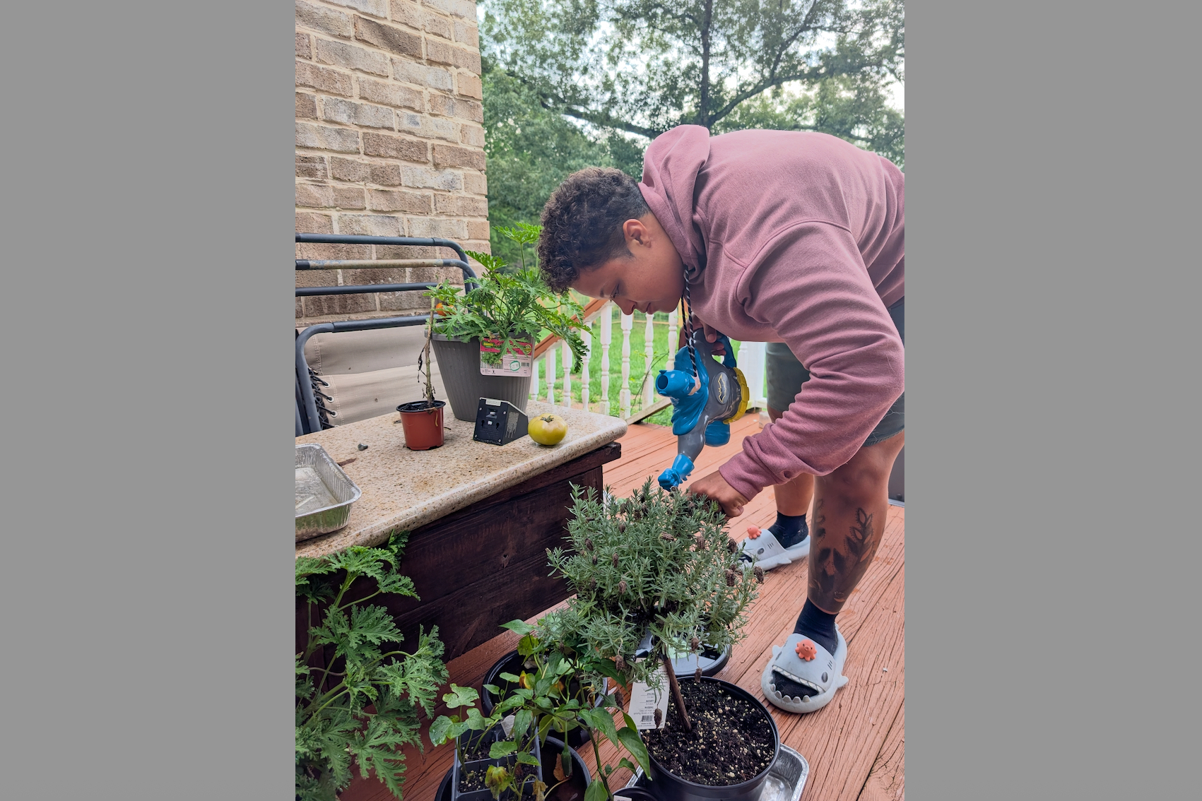 Person in a pink hoodie leaning over a wooden planter on a porch, tending potted plants outdoor.