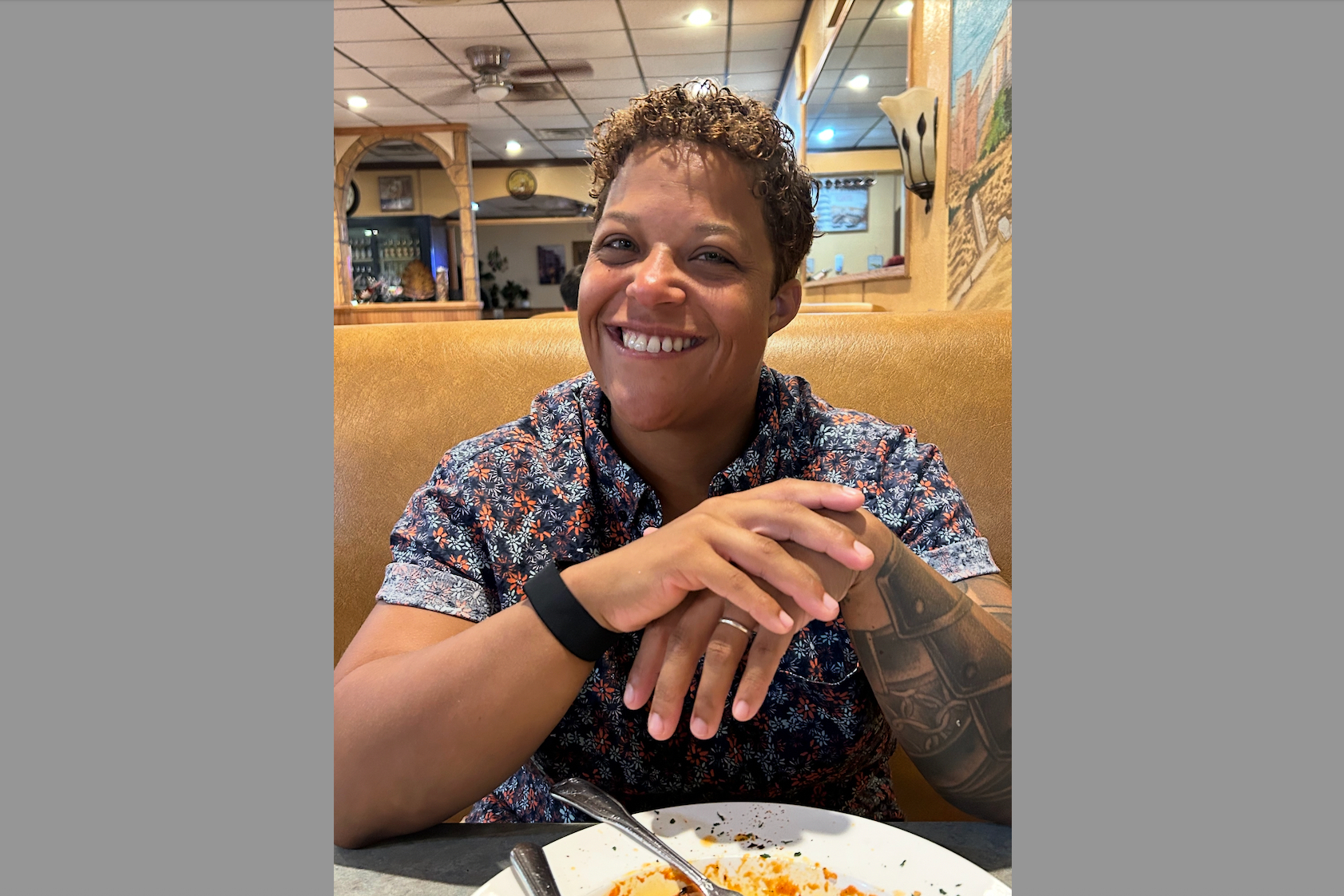 Smiling person with short curly hair sits in a restaurant booth, hands folded on the table, plate in front.