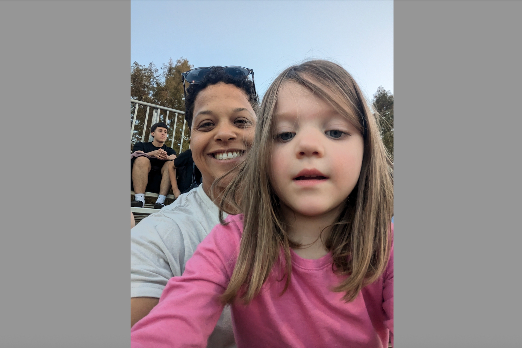 Smiling woman and young girl pose for a selfie outdoors, with a man sitting on bleachers in the background.