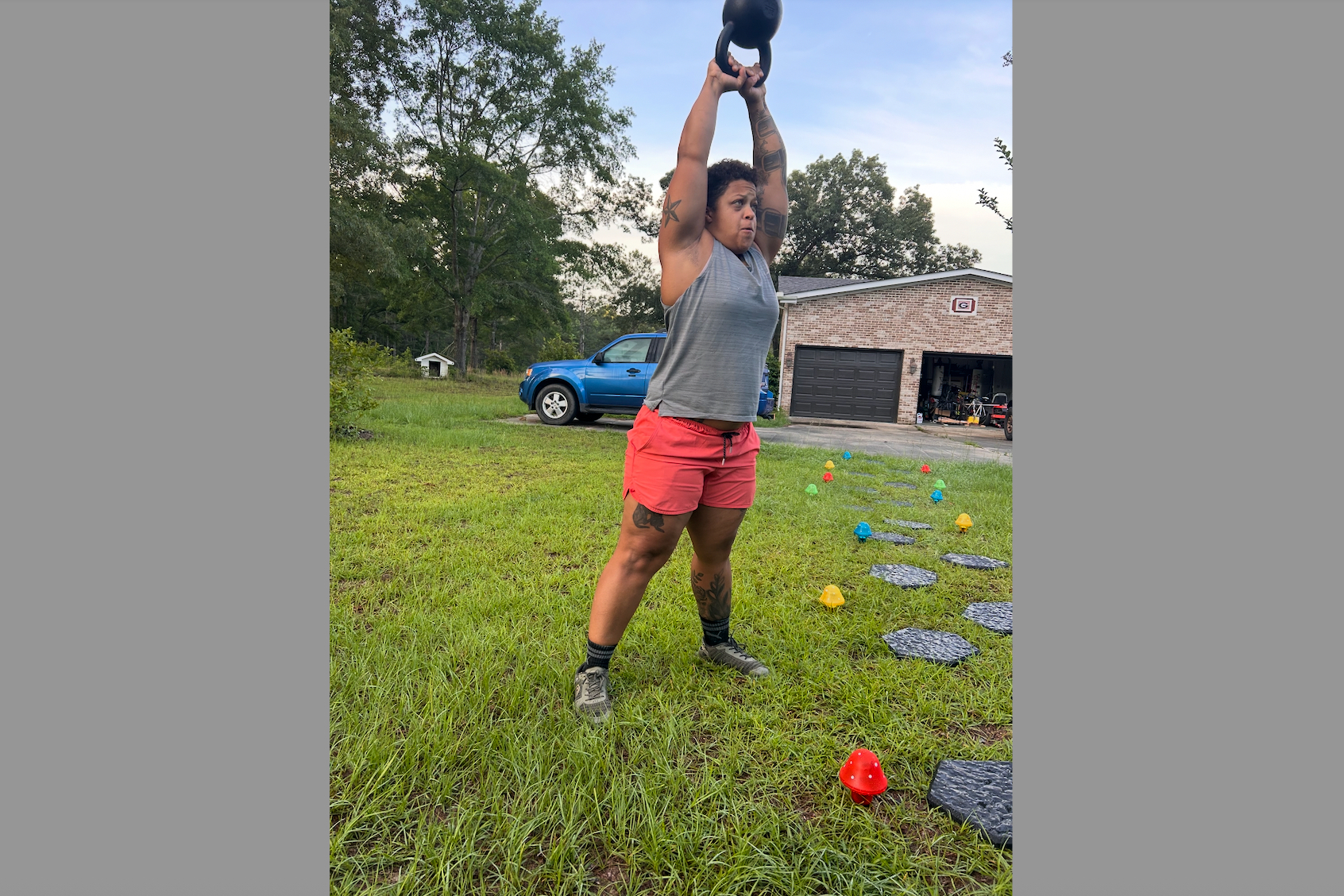 Woman outdoors lifting a kettlebell overhead during a backyard workout, wearing a gray tank top and pink shorts with tattoos visible on both arms, a blue truck and brick garage in the background.