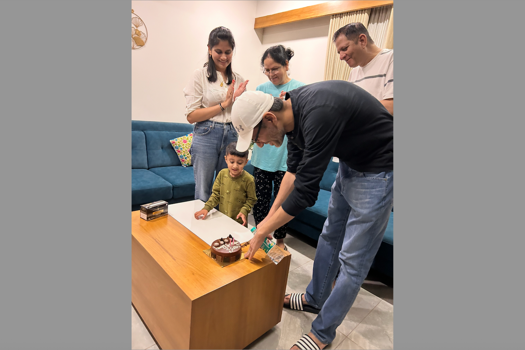 A family gathers to celebrate a birthday: a child stands at a coffee table with a small cake while adults watch and smile.