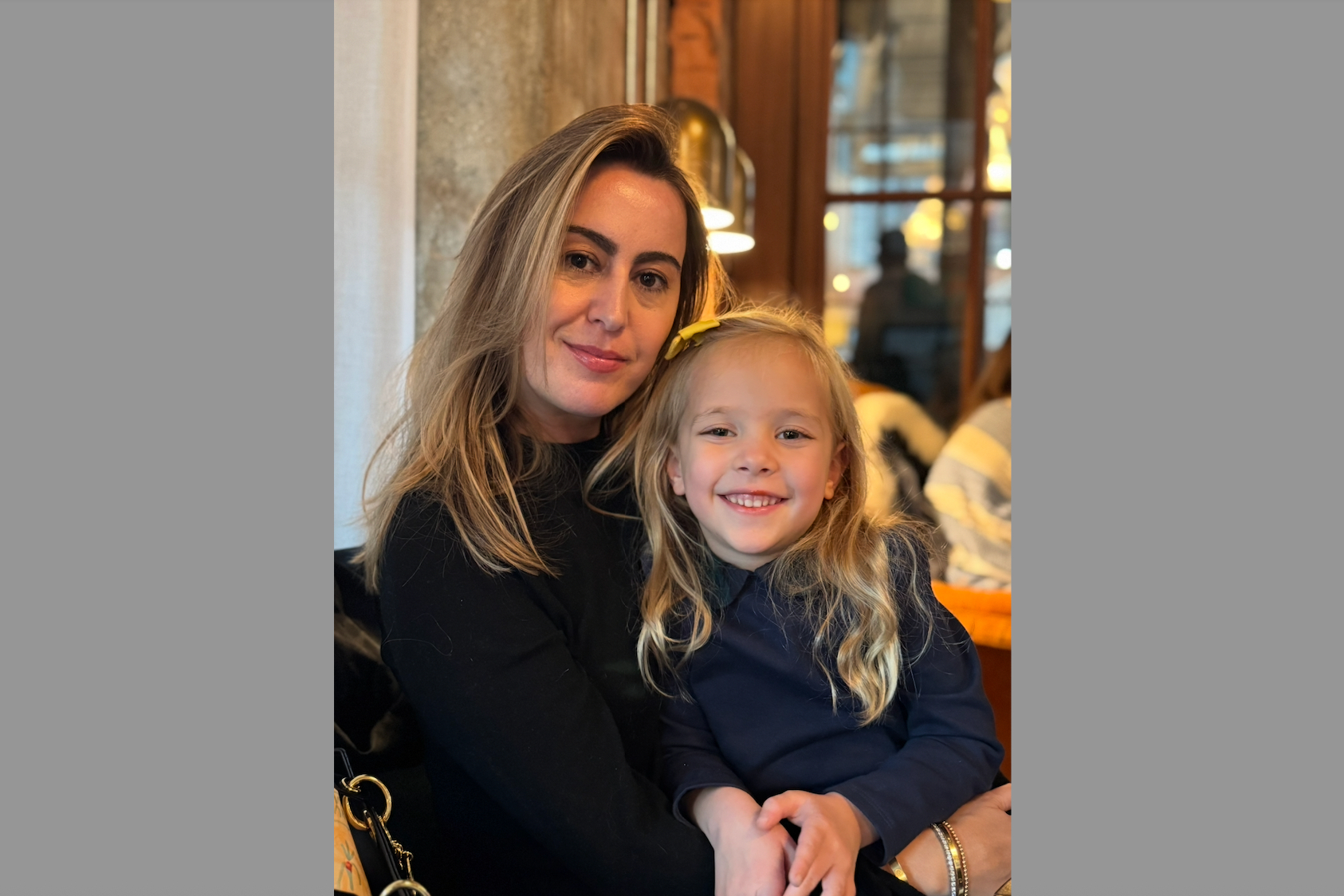 Woman and young girl sit close together, smiling at the camera in a warmly lit cafe.