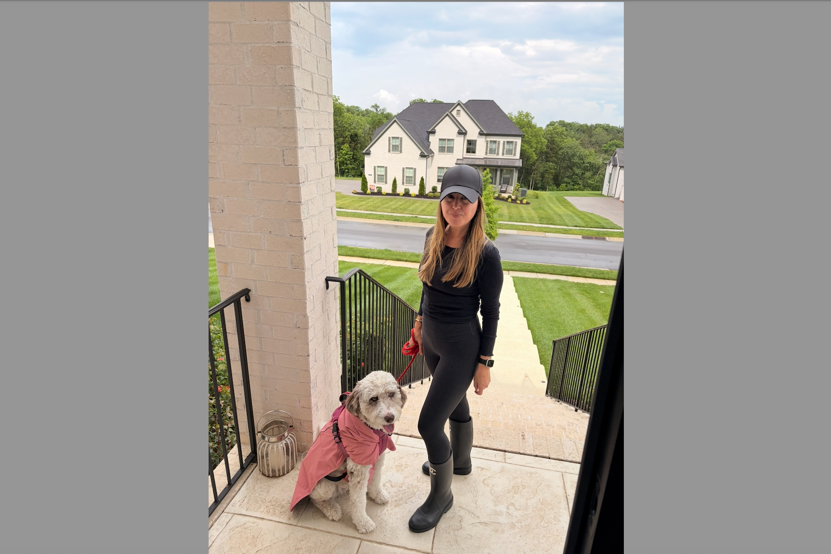 Woman in black athletic wear with cap on porch steps; her dog in a pink raincoat sits beside her.