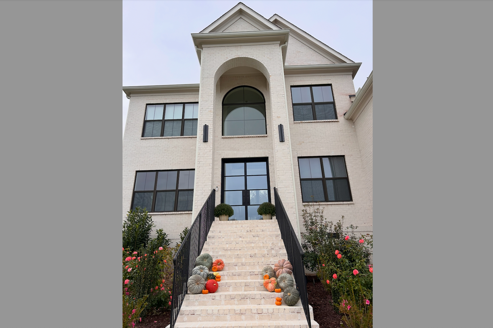 Front stairs of a beige brick townhouse with arched window above the door; pumpkins and gourds line the steps, black railings on both sides.