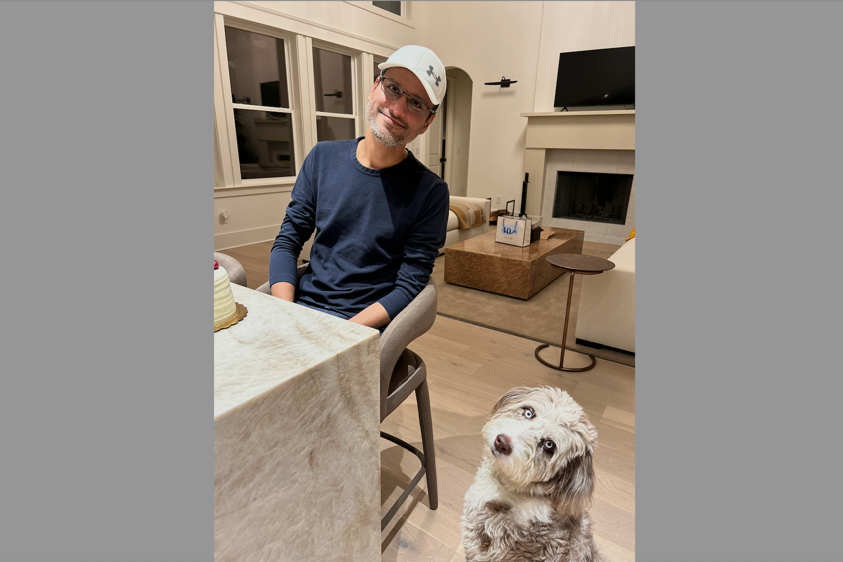 Man wearing a cap and glasses sits at a marble table with a cake, while a fluffy light-colored dog looks up nearby in a living room.
