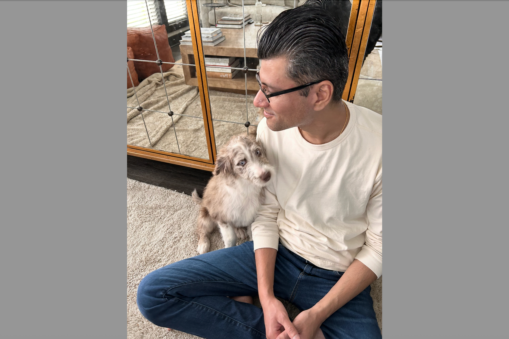 Man in a light sweater sits cross-legged on a beige carpet, looking at a fluffy puppy beside him indoors.