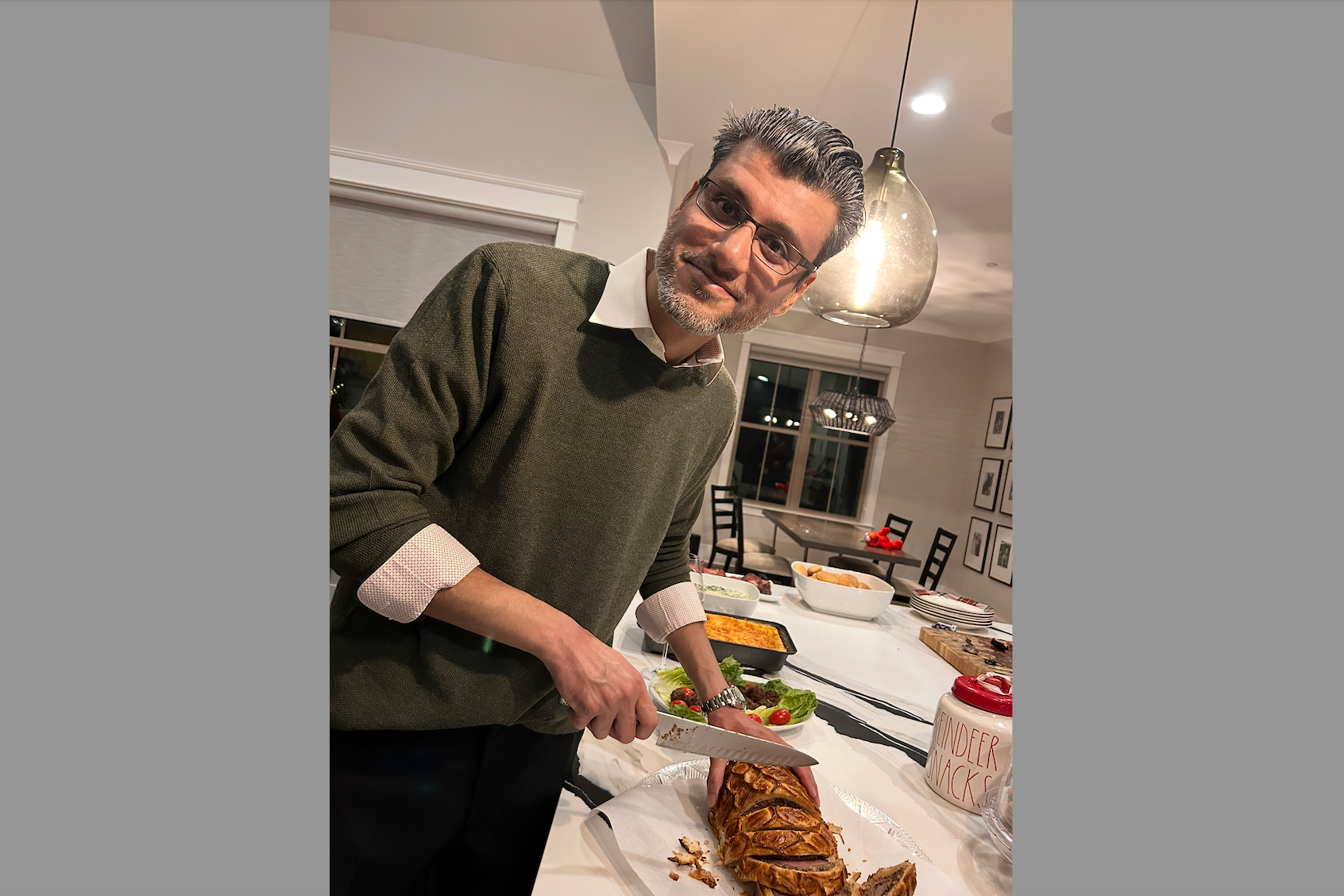 Man in a green sweater slices a loaf of bread at a kitchen island during a gathering, with bowls and plates in the background.