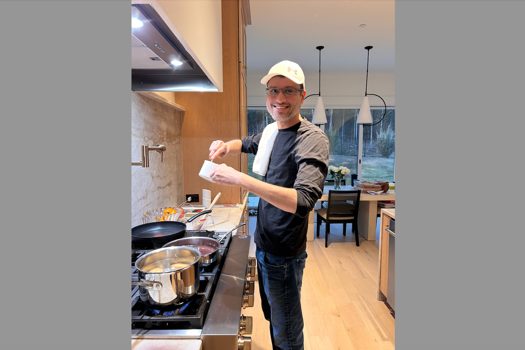Smiling man wearing a white cap and glasses standing at a kitchen stove, stirring a pot.