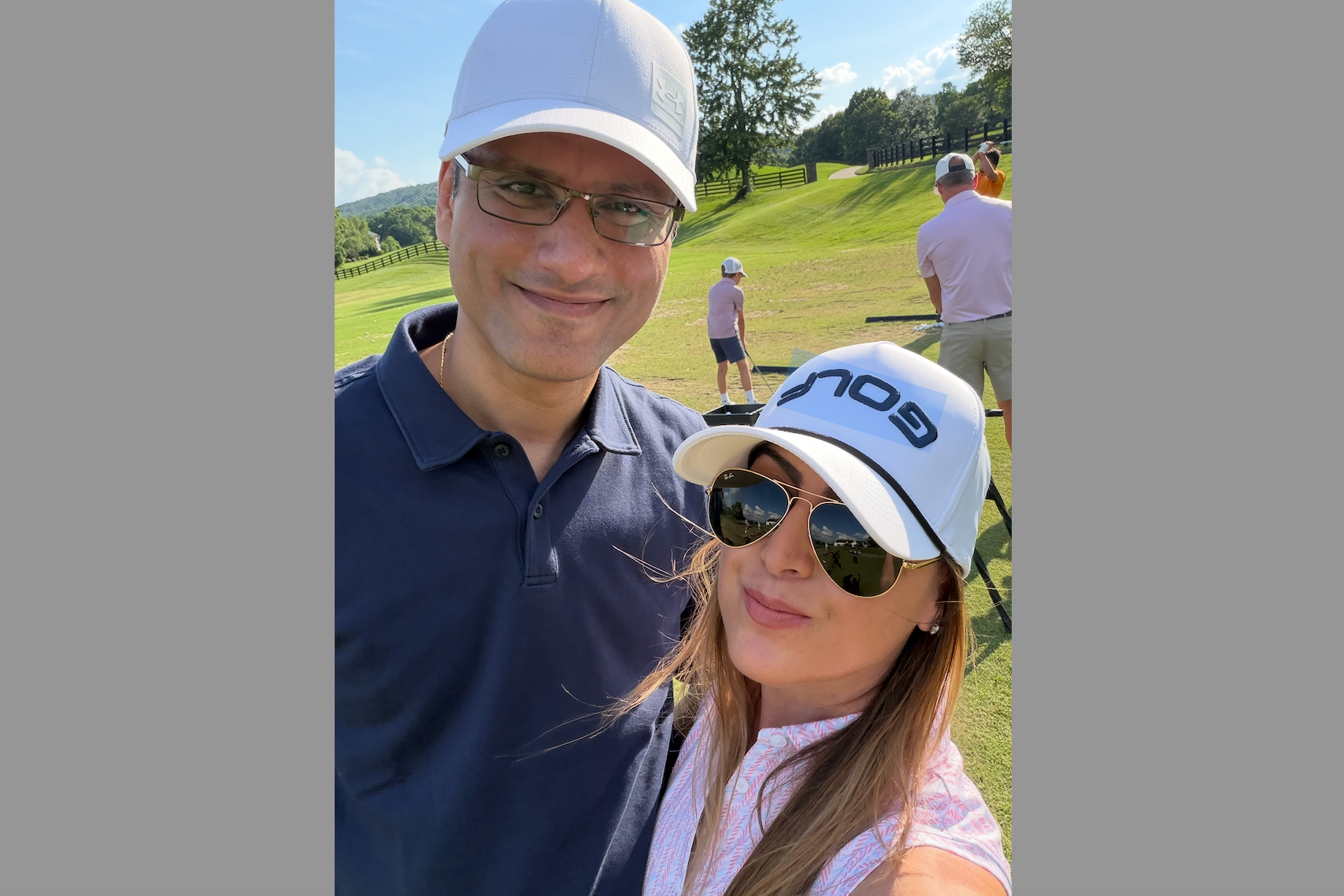 Couple taking a selfie on a sunny golf course; man in a navy polo and white cap, woman in white cap and sunglasses in foreground.