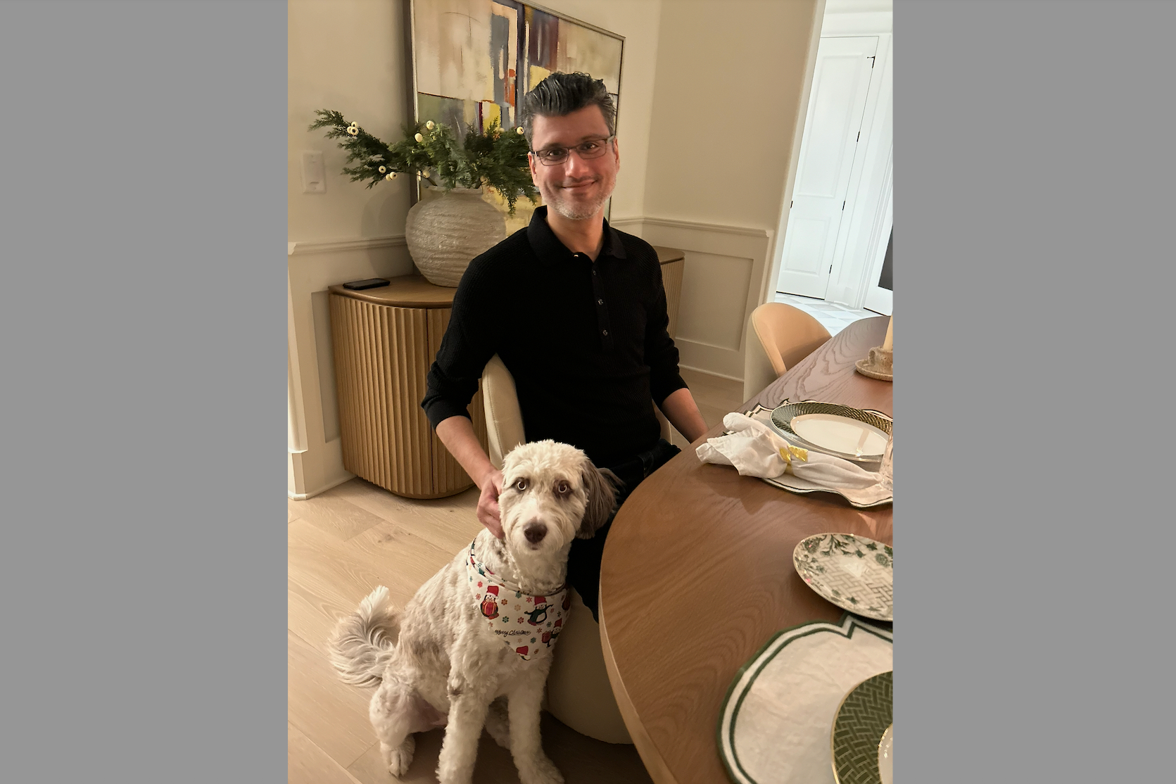 A man in a black polo sits at a dining table with a white fluffy dog wearing a holiday bandana beside him in a home setting.