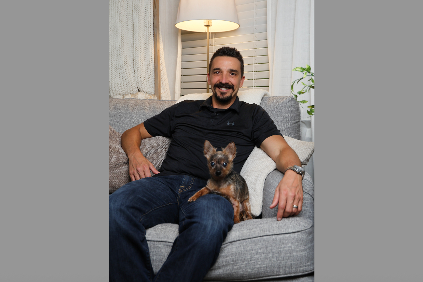 Smiling man in a black polo shirt sits on a gray sofa with a small brown and black terrier on his lap in a cozy living room.