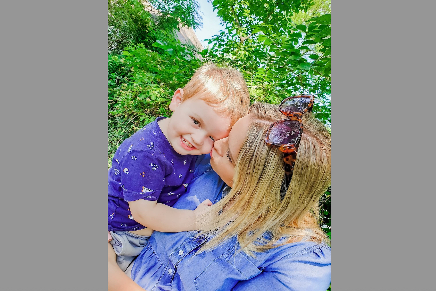 Mother and young child smiling and cuddling outdoors among green foliage.