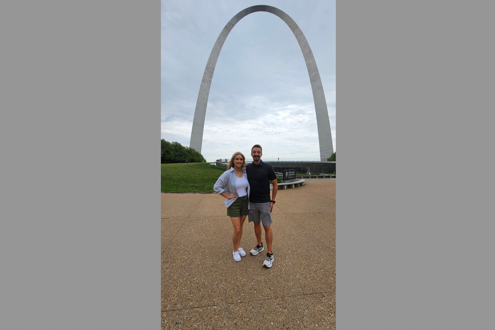 Couple posing in front of the Gateway Arch in St. Louis, smiling at the camera on a cloudy day.