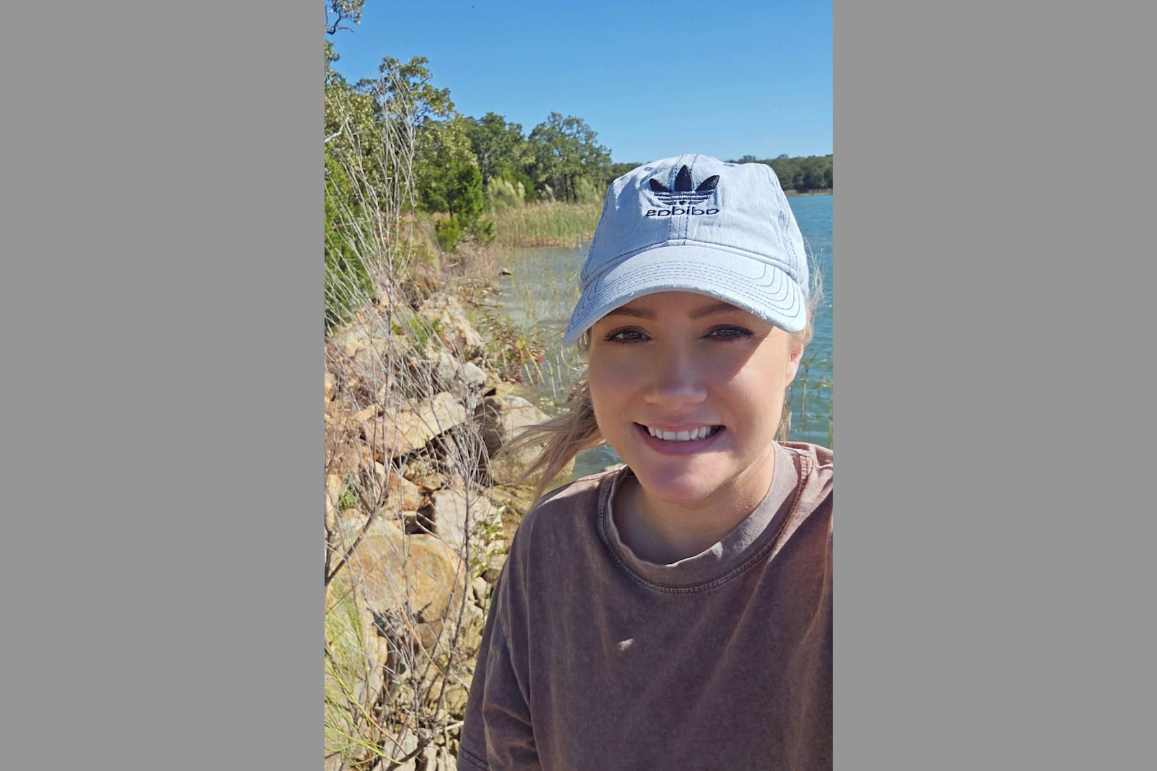 Woman wearing a light blue Adidas cap smiles at the camera by a lakeside with rocks and trees nearby, on a sunny day.