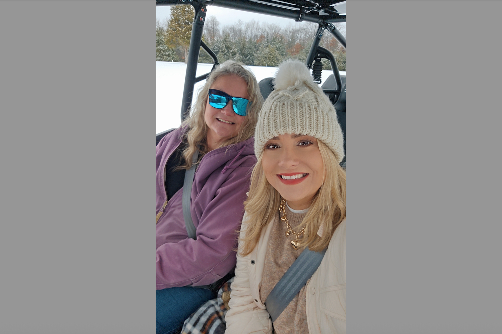 Two smiling women in winter gear pose for a selfie inside a snowmobile, one in a beige pom-pom beanie and the other in blue mirrored sunglasses.