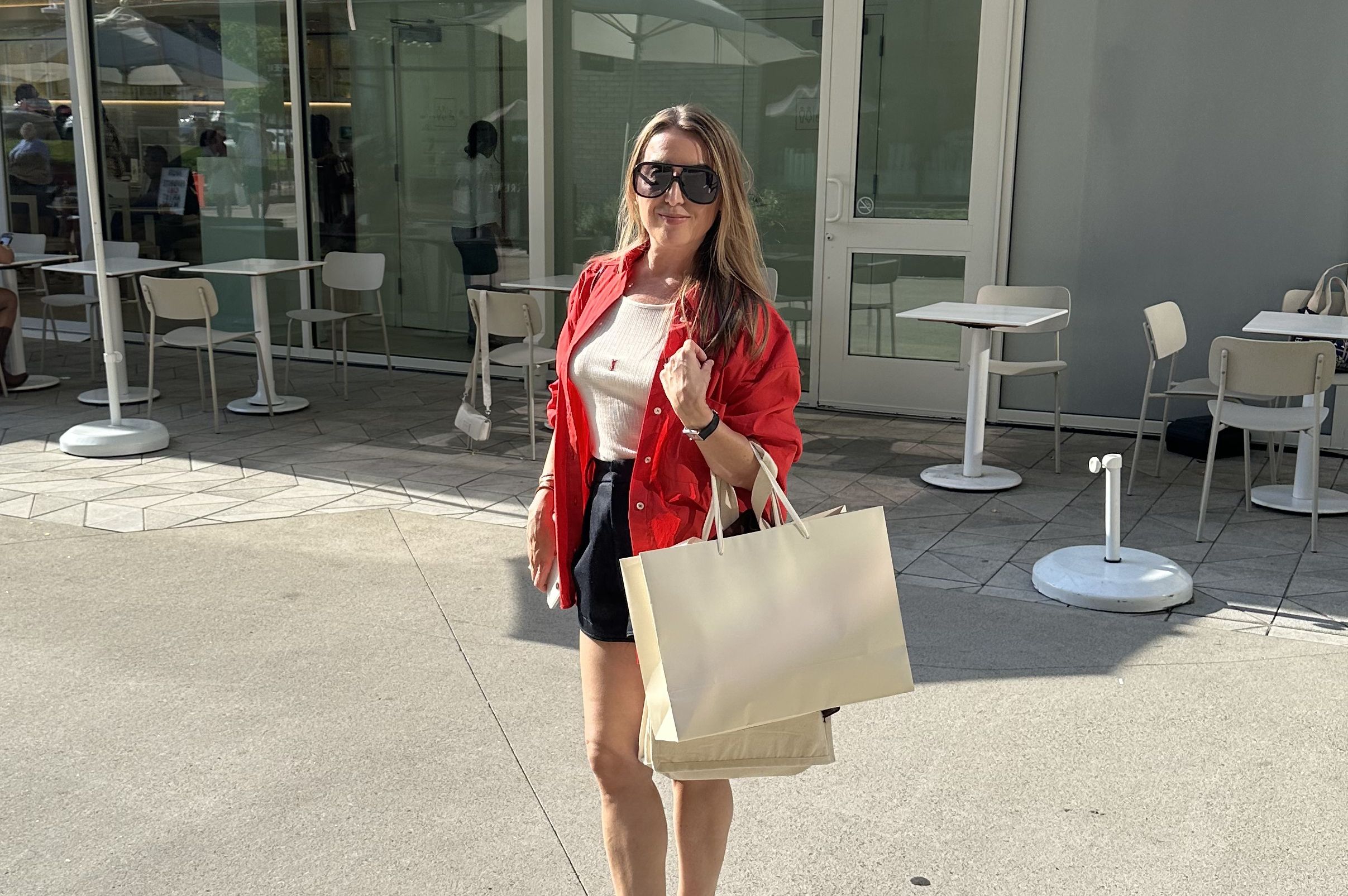Woman standing outside a cafe with beige shopping bags, wearing a red shirt, white top, black shorts, and sunglasses, posing for a photo.