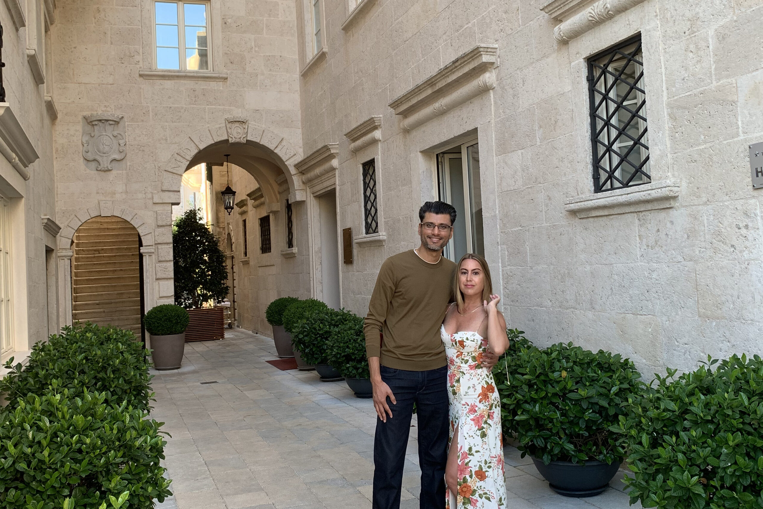 Smiling couple standing in a beige stone courtyard with arches and potted shrubs behind them.