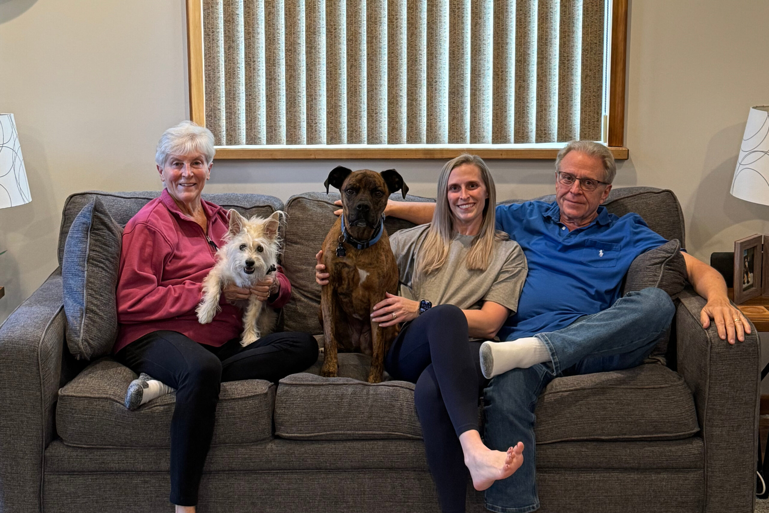 A casually dressed family of four sitting on a sofa with two dogs: a small white fluffy dog on the woman's lap (left) and a brown dog beside the younger woman (center). The older man (right) wears a blue shirt, and a neutral living room backdrop is visible behind them.