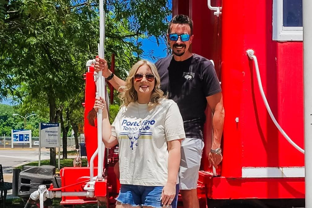 Smiling woman in a Portofino Tennis Club tee and a man in a black shirt stand on a bright red amusement-ride frame outdoors, both wearing sunglasses.