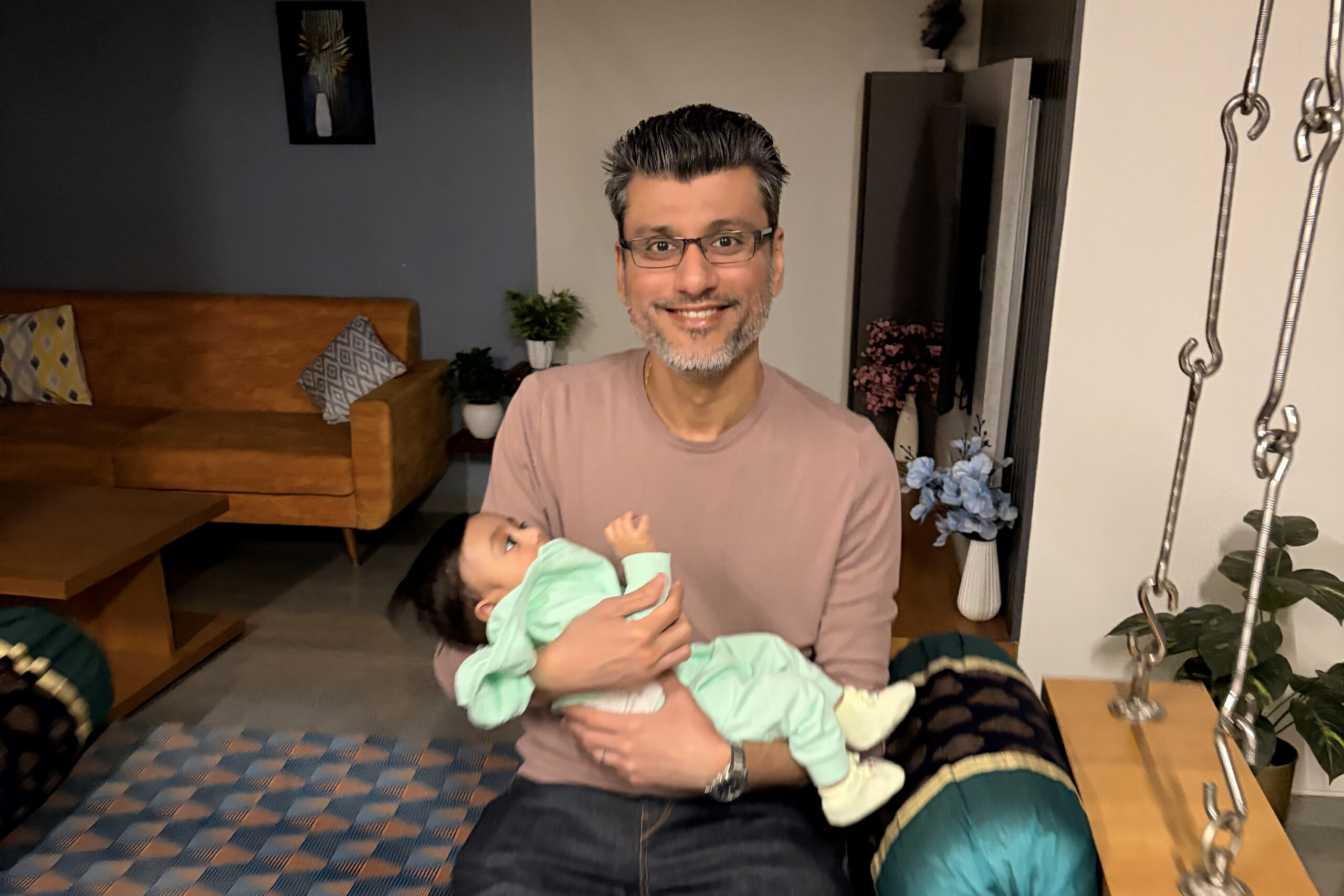 Man with glasses smiles while holding a baby in a cozy living room.