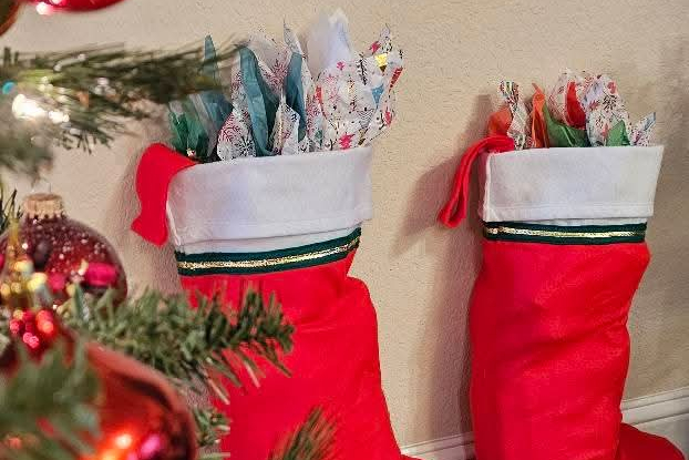 Two red Christmas stockings with white cuffs and green-gold trim, filled with tissue-wrapped gifts, hanging by a wall next to a tree.