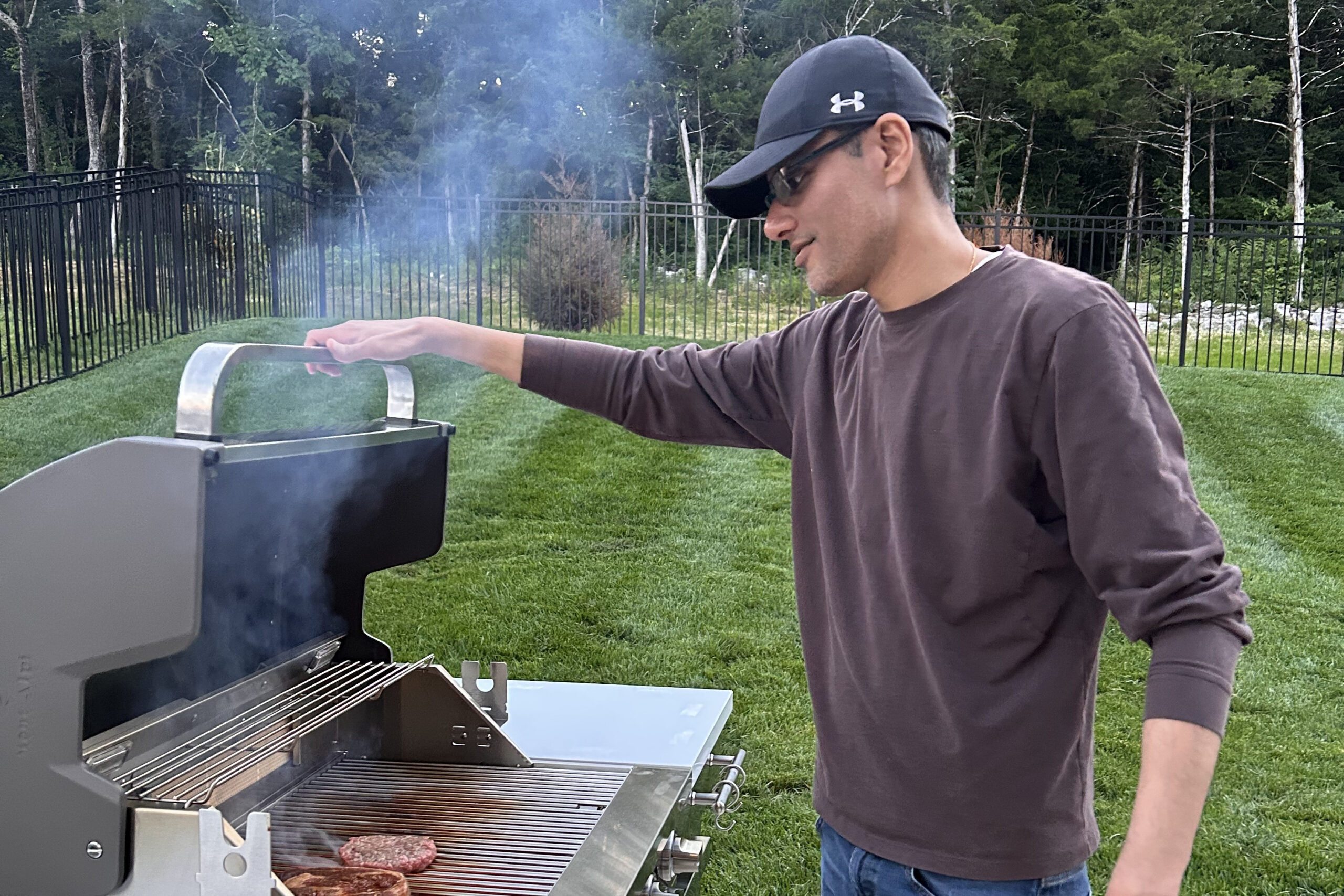 Man in a dark long-sleeve shirt and cap grilling burgers at an outdoor barbecue in a grassy backyard.