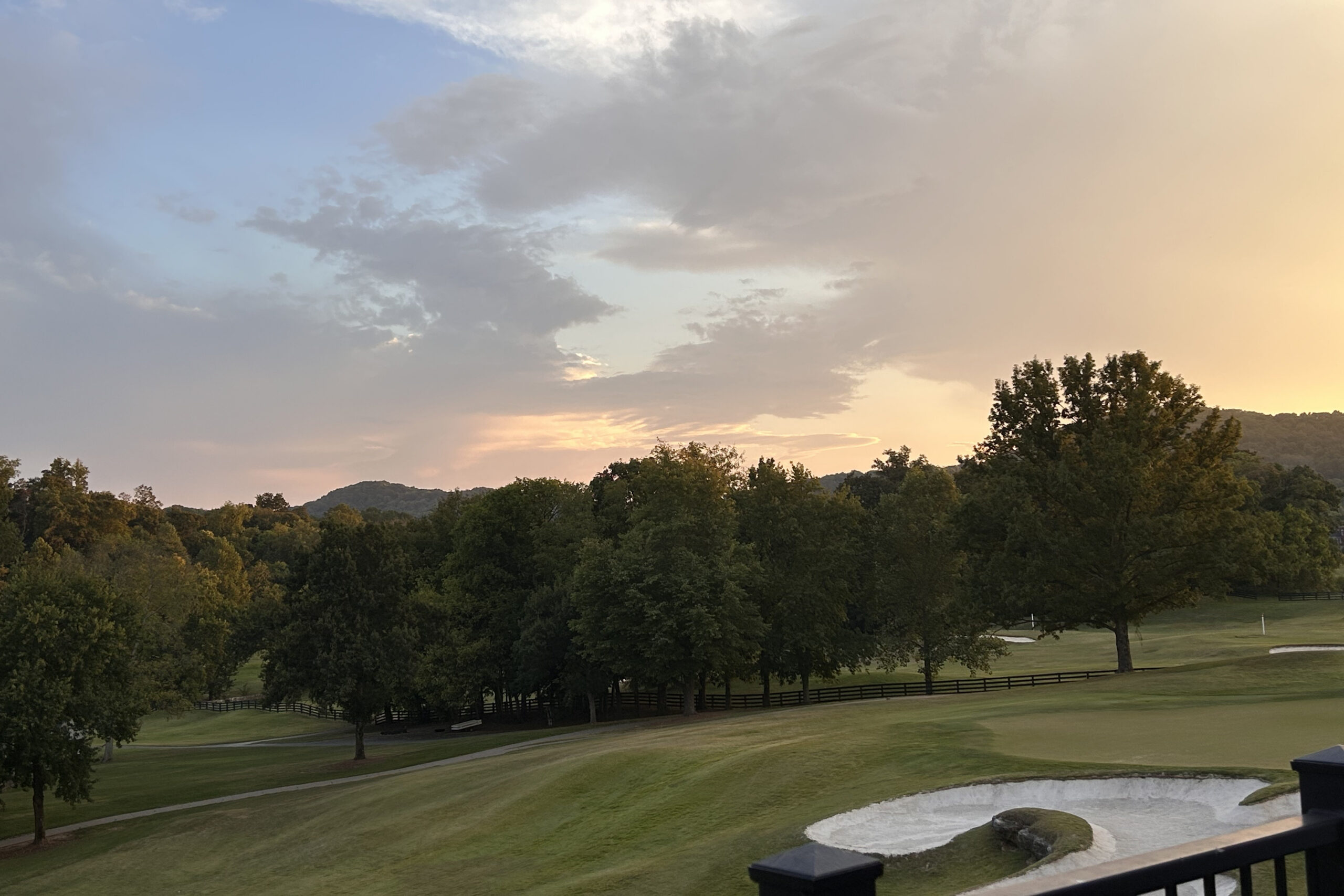 Sunset over a golf course: green fairways, a line of trees, hills in the distance, and a sandy bunker in the foreground.