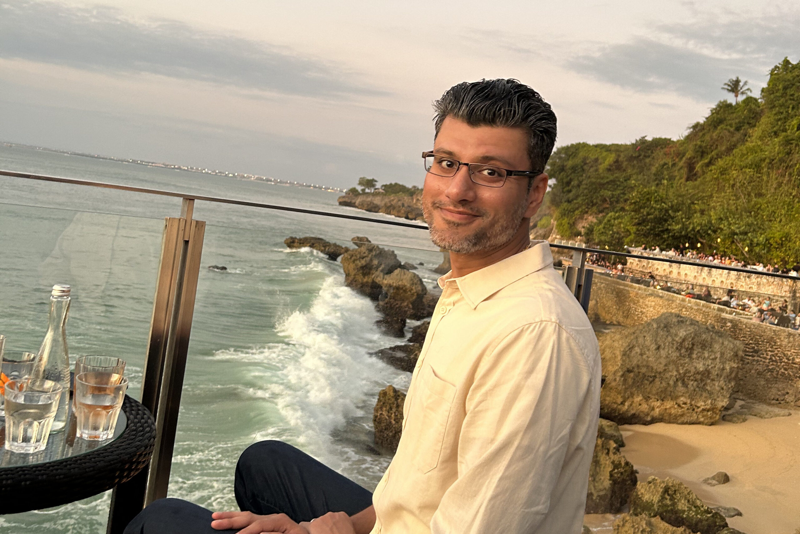 Man wearing glasses and a light beige shirt sits at an outdoor seaside restaurant, with the ocean and rocky coastline behind him.