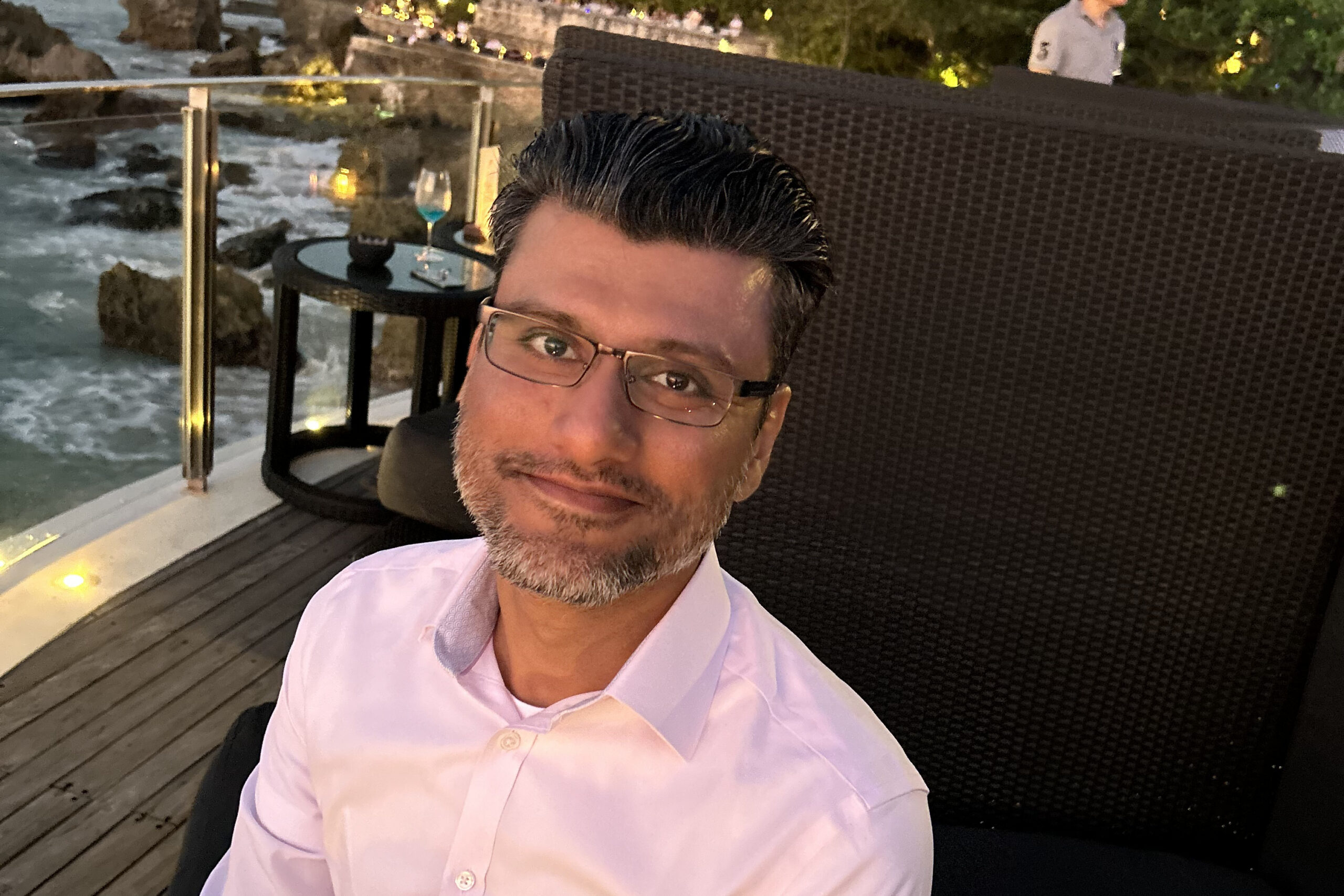 Selfie of a man in a pink shirt and glasses seated on a wicker lounge chair on a wooden deck by the water at dusk.