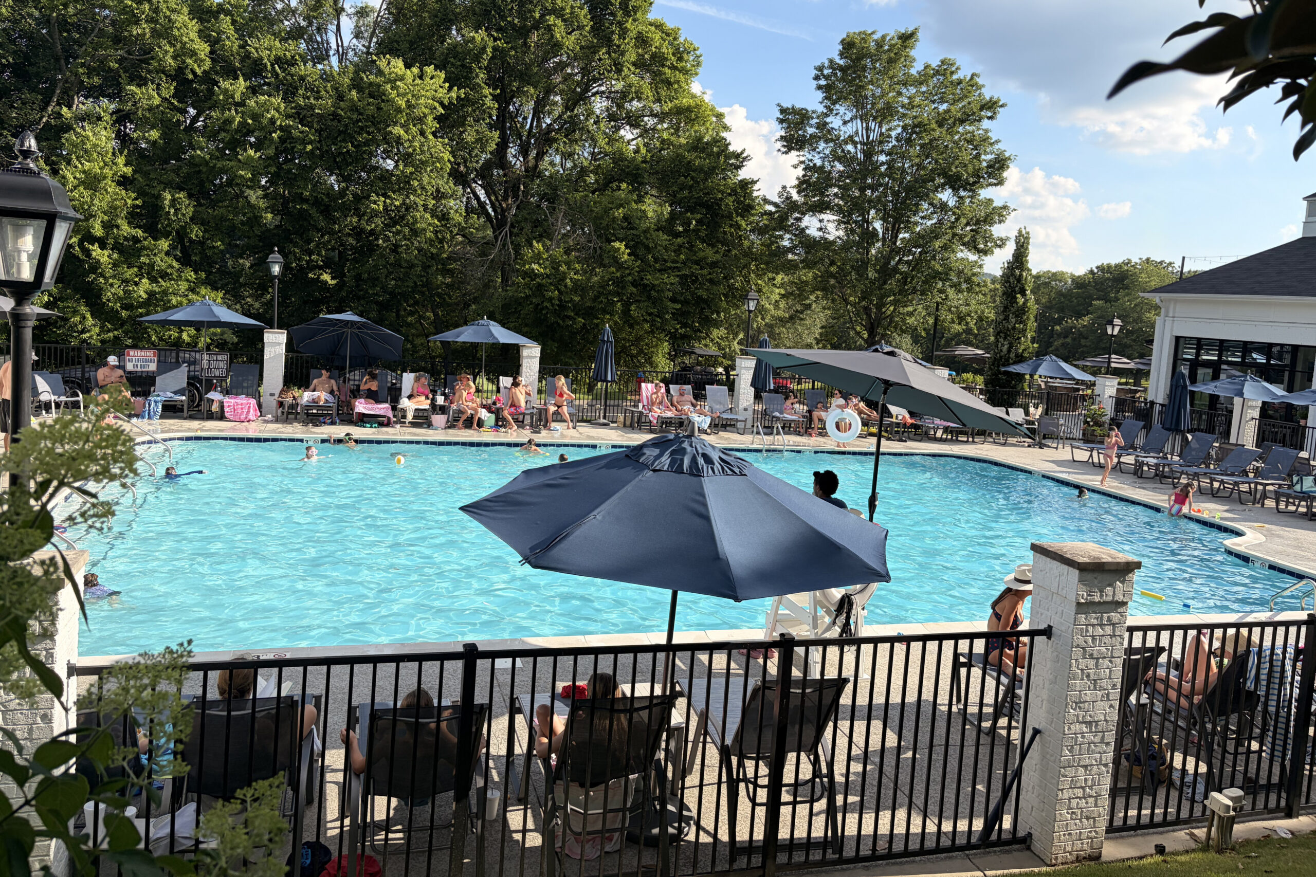Poolside at a sunny resort with numerous lounge chairs, umbrellas, and guests relaxing by the water behind a black fence.