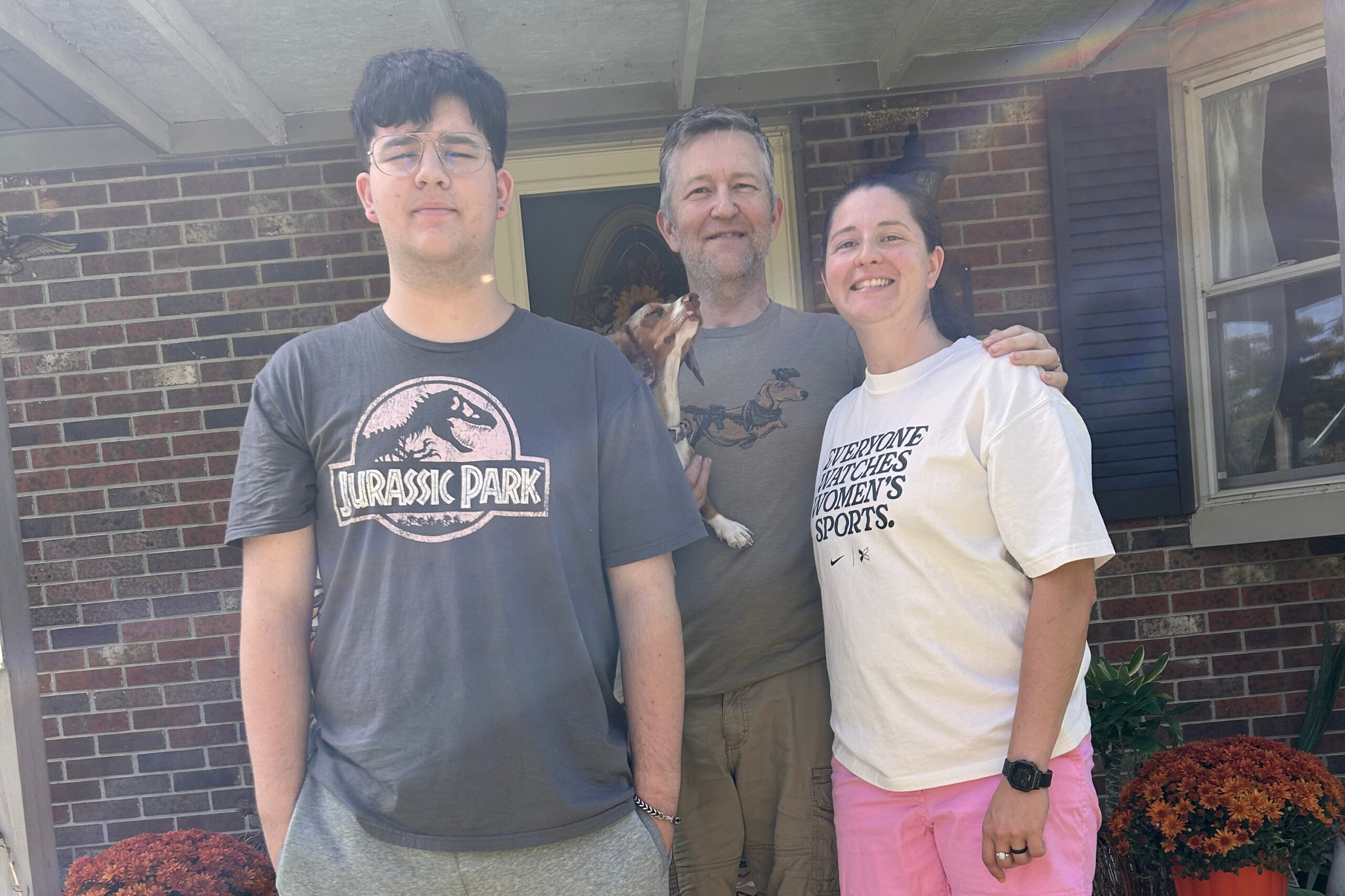 Three adults pose on a front porch with a small dog between them, in front of a brick house.