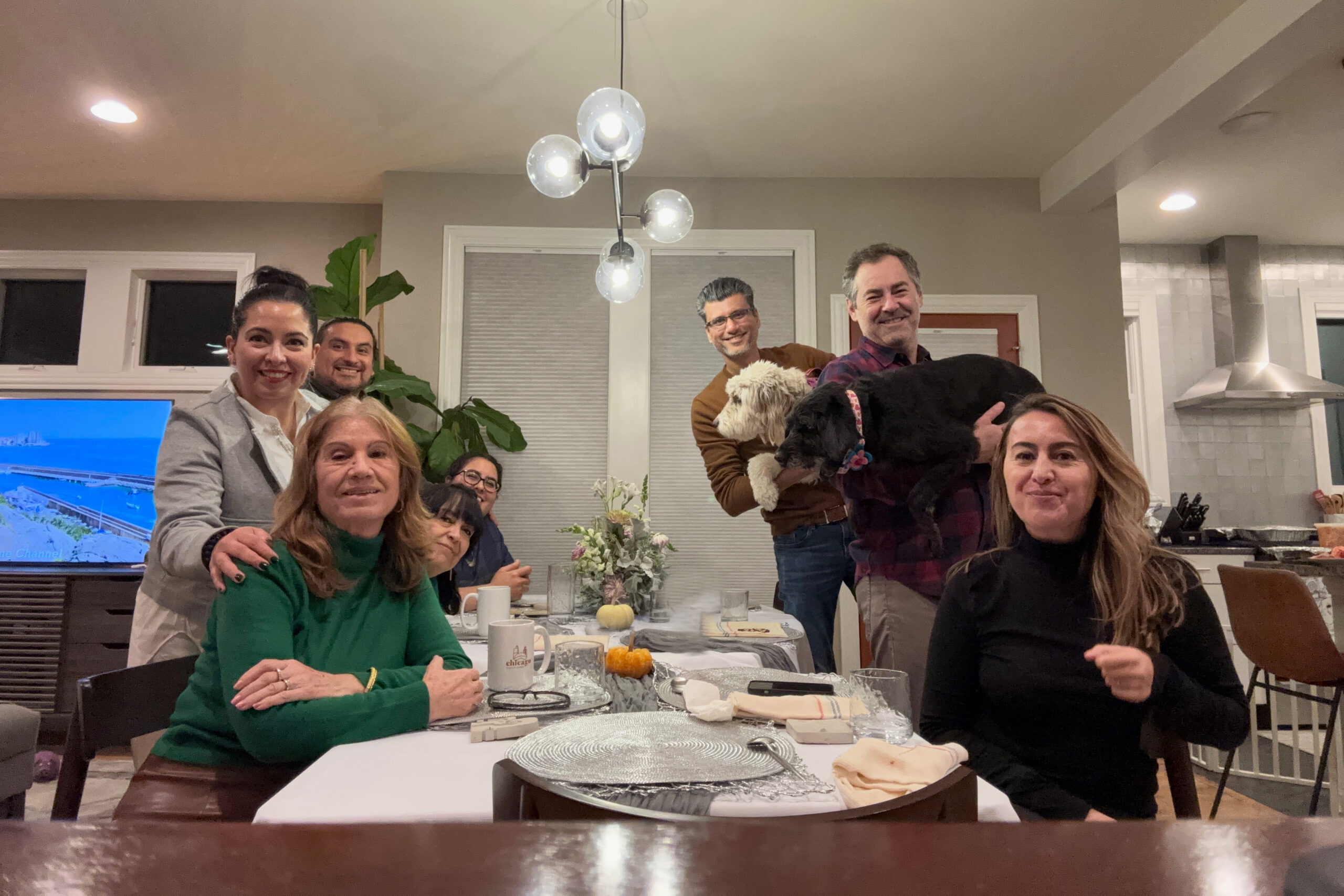Group of people gathered around a dining table in a bright kitchen, smiling at the camera, with a dog perched on a lap and pumpkins on the table.