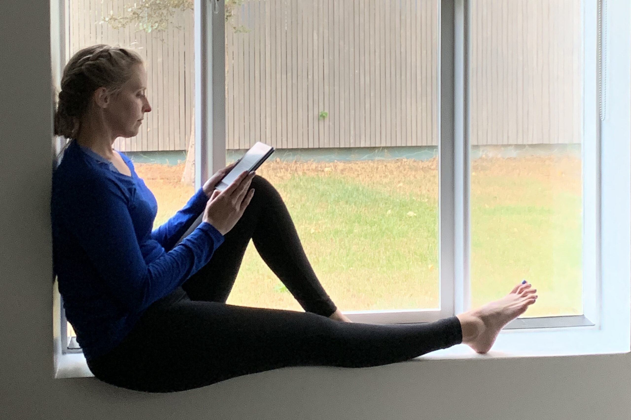 Woman in a blue top sits on a windowsill reading a tablet with legs extended on the ledge.
