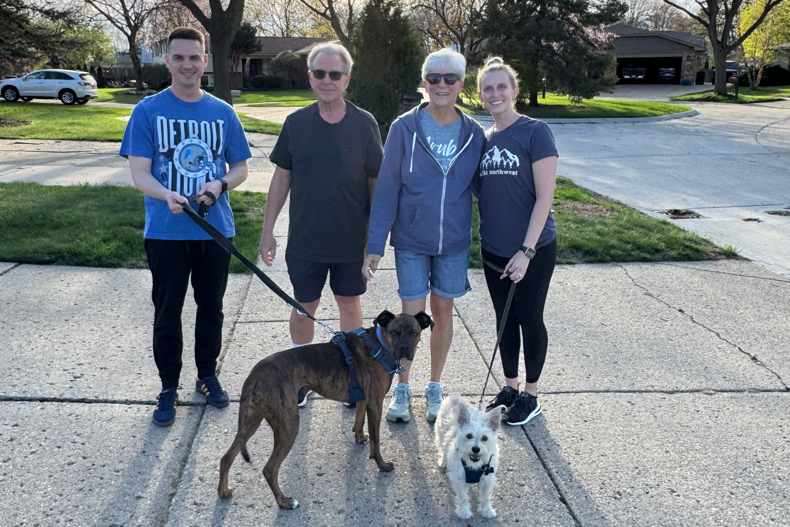 Four people stand together on a suburban street with two dogs in the foreground, smiling at the camera on a sunny day.
