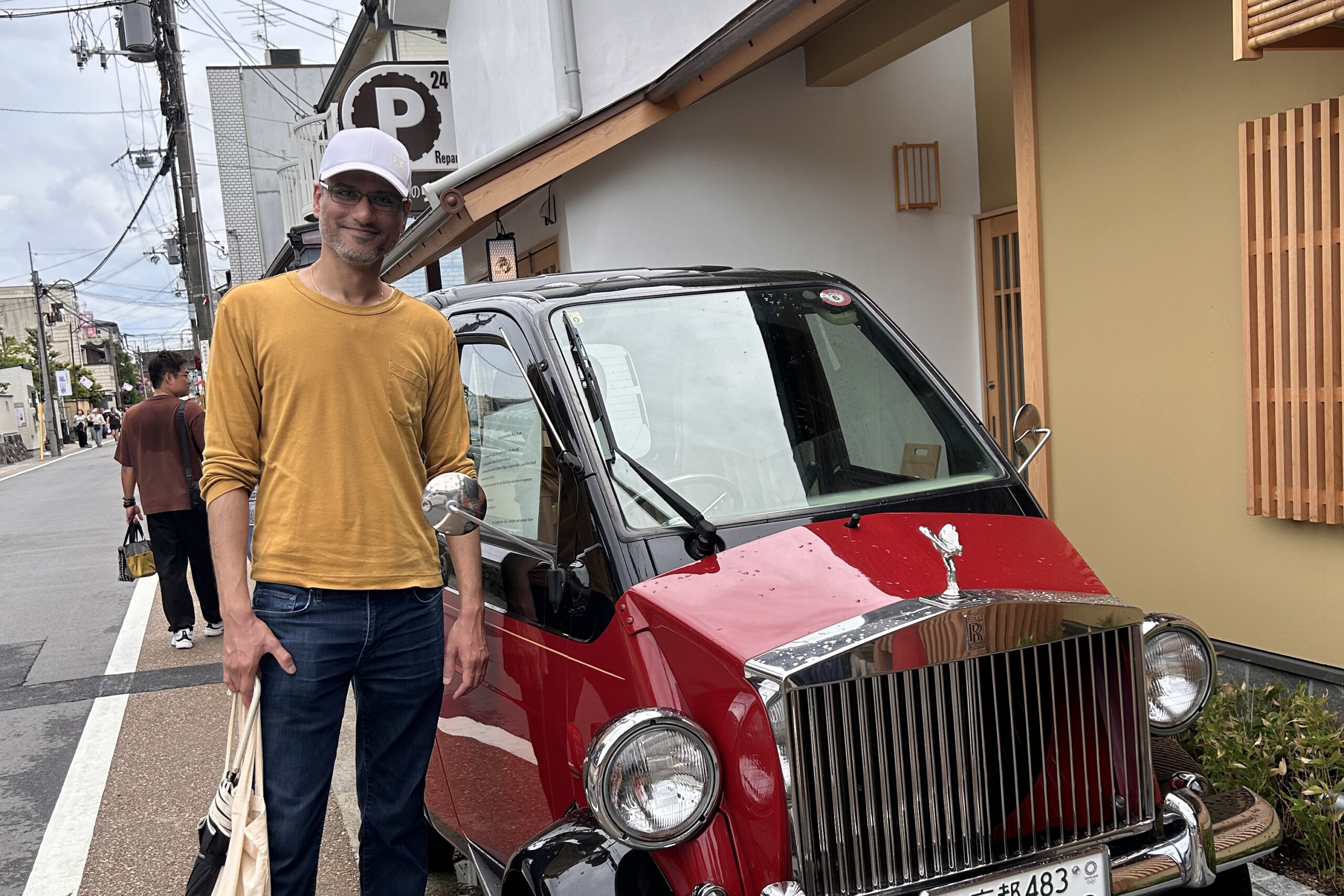 Man in a mustard long-sleeve shirt and white cap stands beside a red vintage Rolls-Royce on a city street, smiling at the camera.