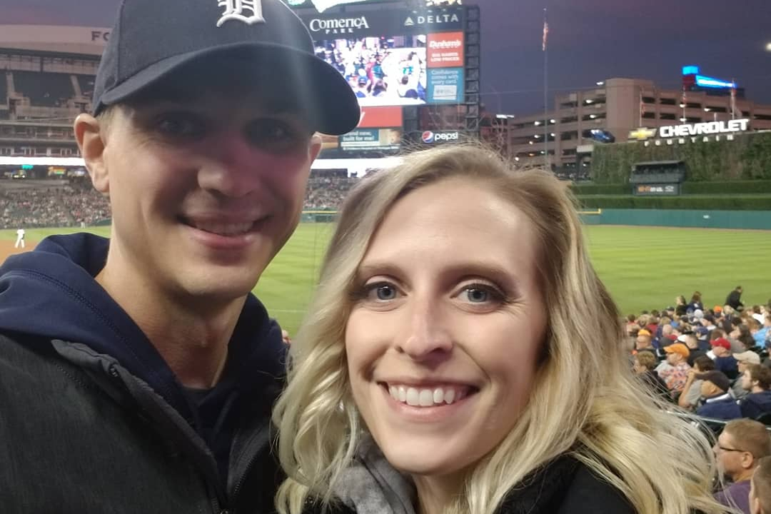 Smiling couple at a baseball stadium during a night game, with the field and scoreboard behind them.