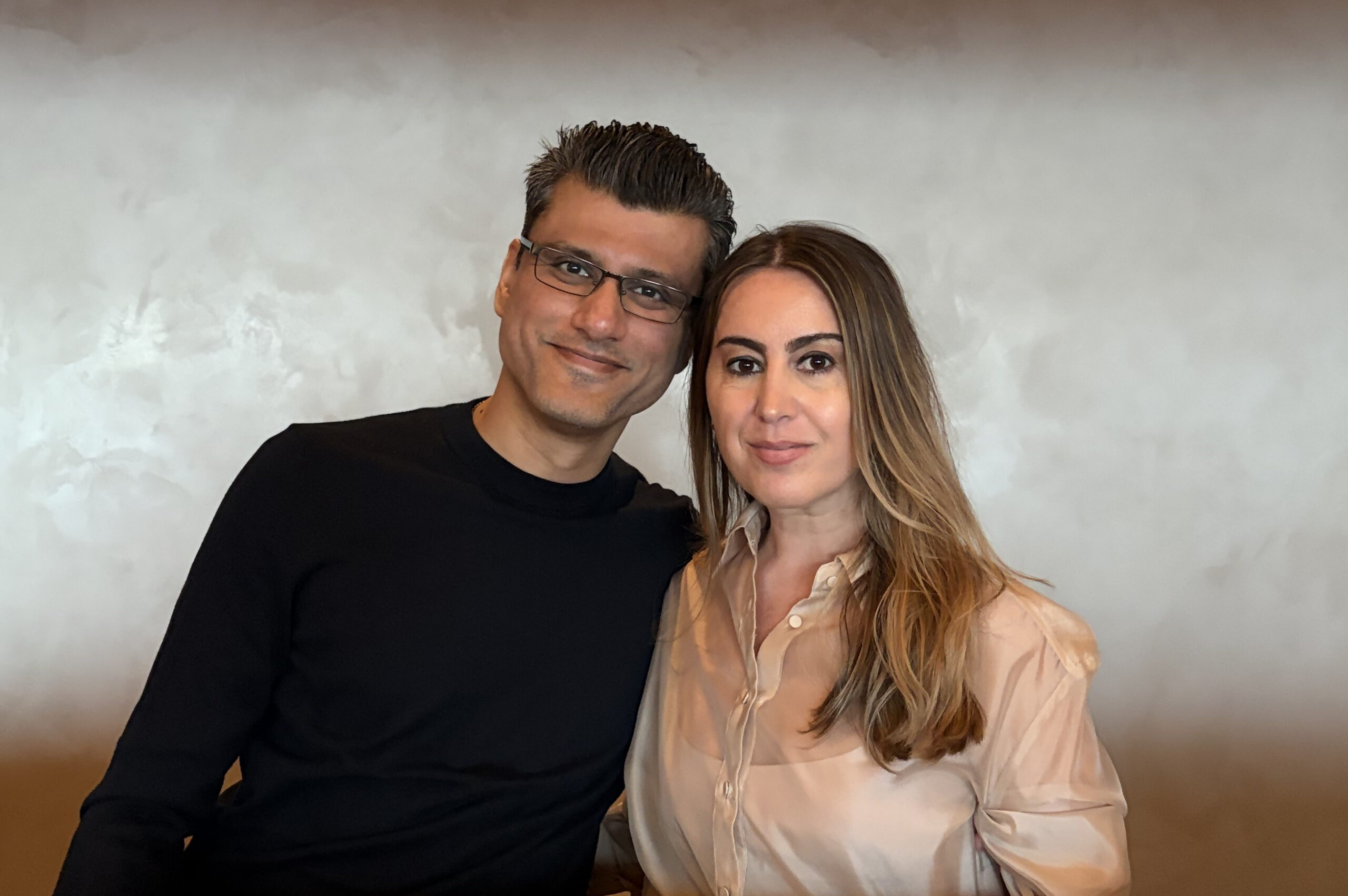 Smiling man in a black shirt and a smiling woman in a light blouse pose together against a textured gray wall.