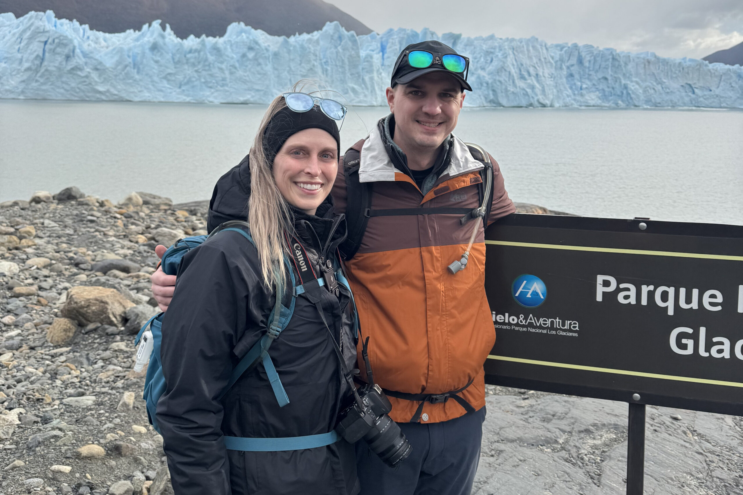 Smiling couple in outdoor jackets posing on a rocky shore with a large blue glacier behind them.