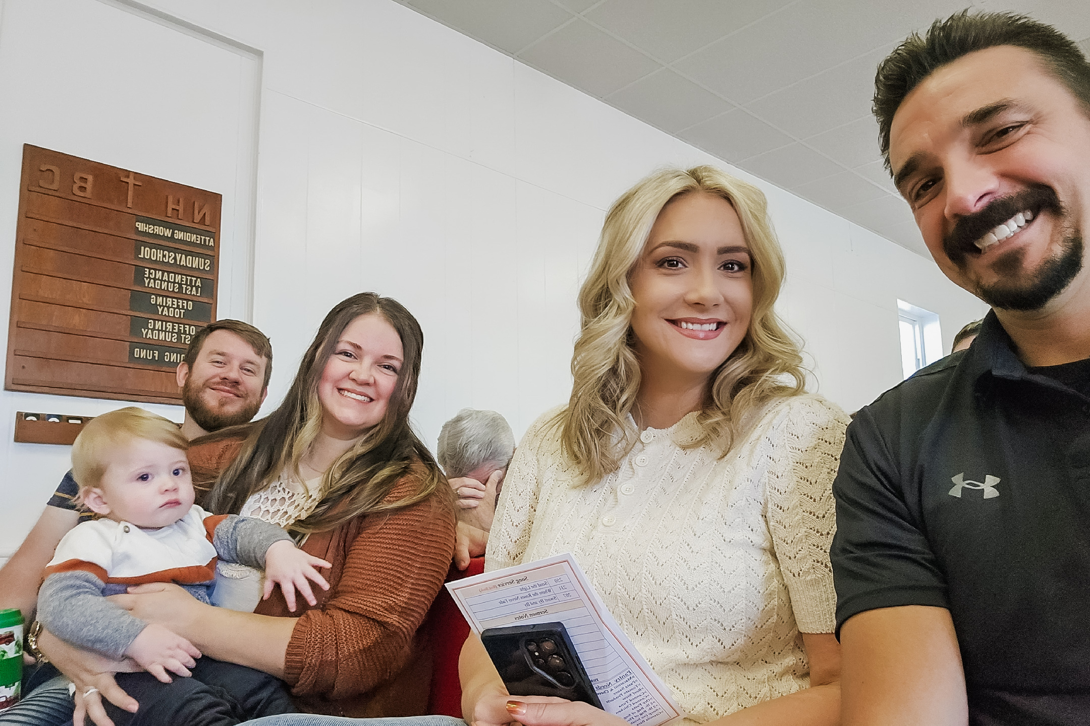 Group selfie with several adults and a baby smiling indoors, white walls behind them.