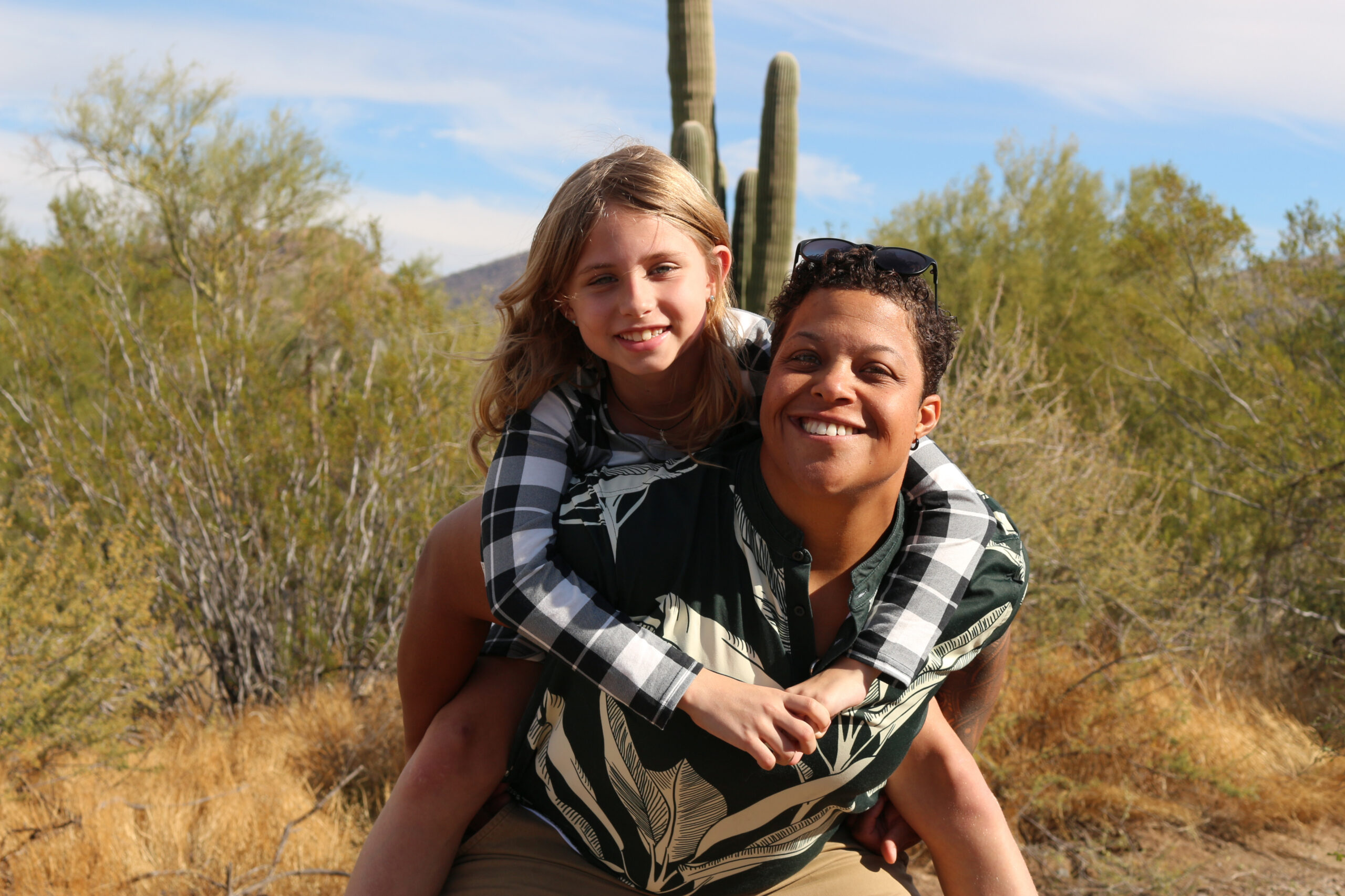 Two people are outdoors in a desert, one giving the other a piggyback ride while smiling at the camera, with a tall cactus and scrub behind them.
