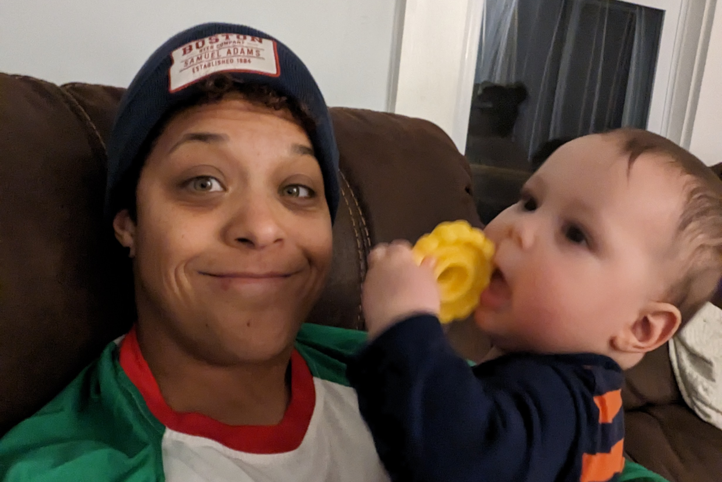 Woman wearing a dark knit hat sits on a brown couch, smiling as a young child shares a yellow snack nearby — close-up.