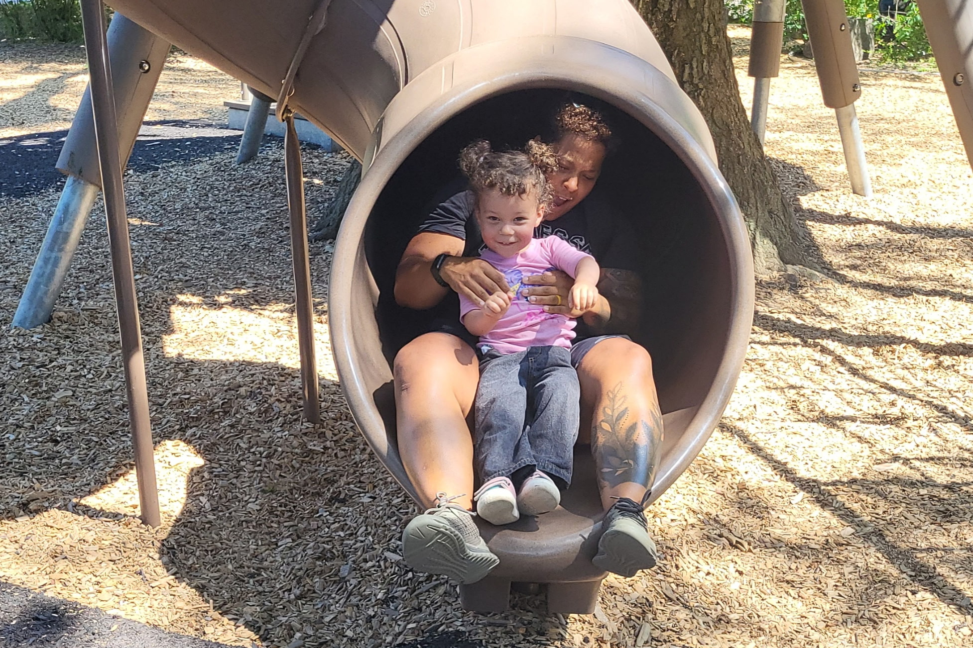 Adult with tattooed legs sits with a child at the end of a large playground tunnel slide; both look toward the camera; wood chips on the ground and trees in the background.