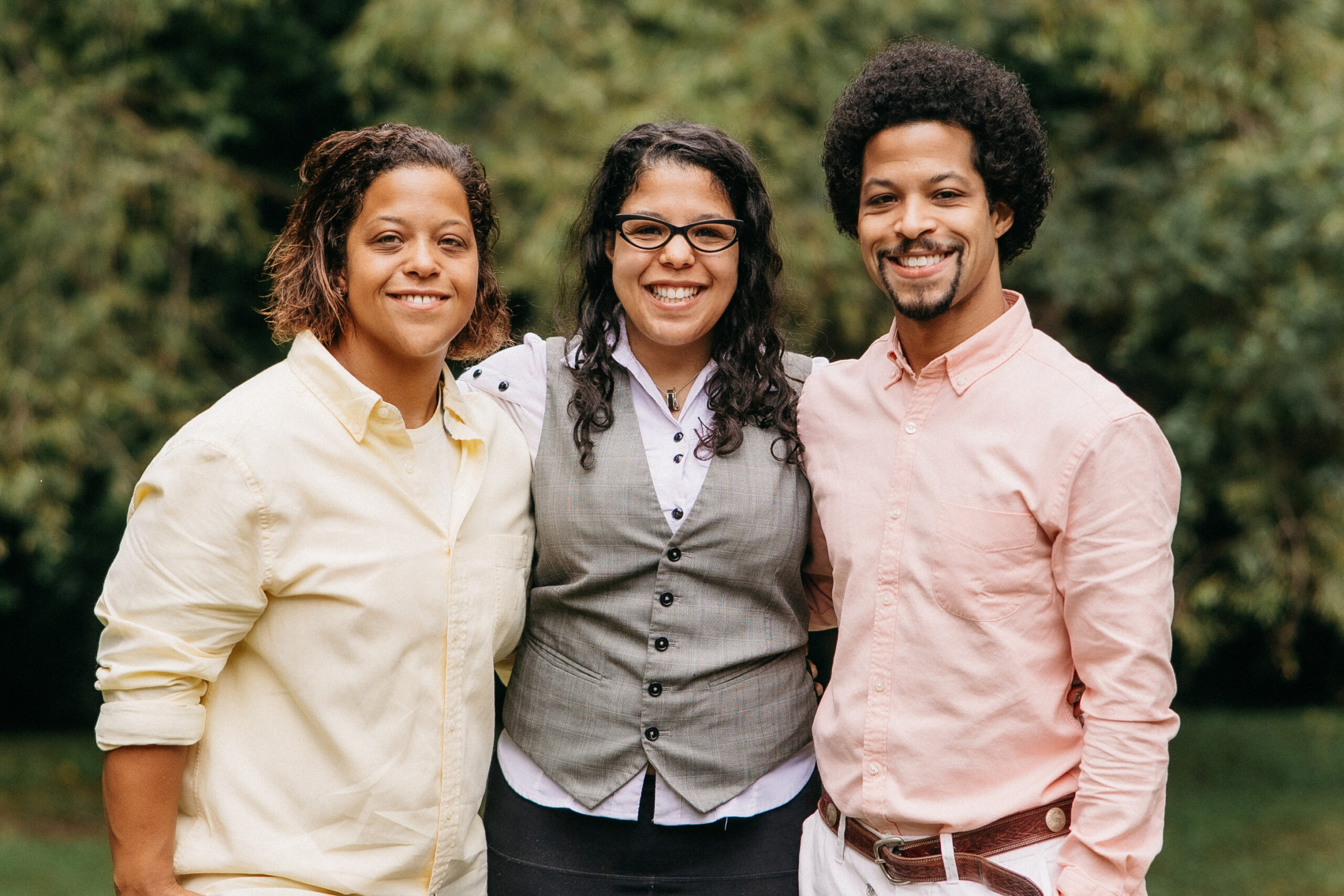 Three friends stand close together outdoors, smiling at the camera; left in a pale yellow shirt, center in a gray vest with glasses, right in a pink shirt.