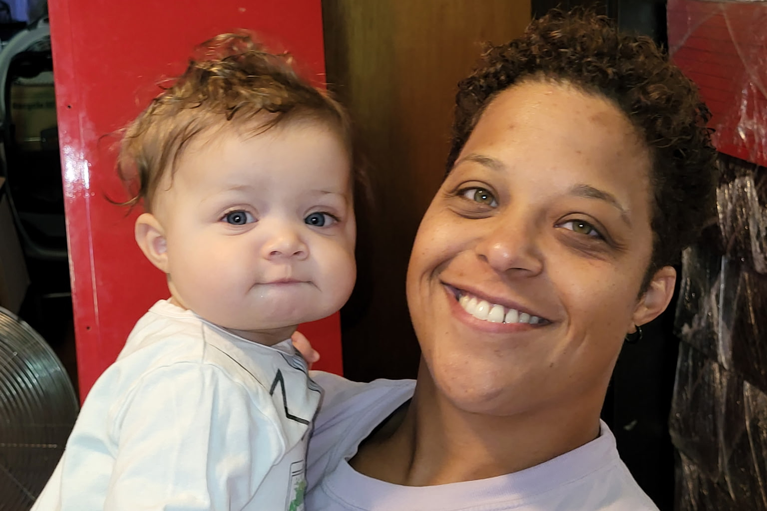 Woman with short curly hair smiling while holding a baby toward the camera. They’re indoors against a red background or pillar.